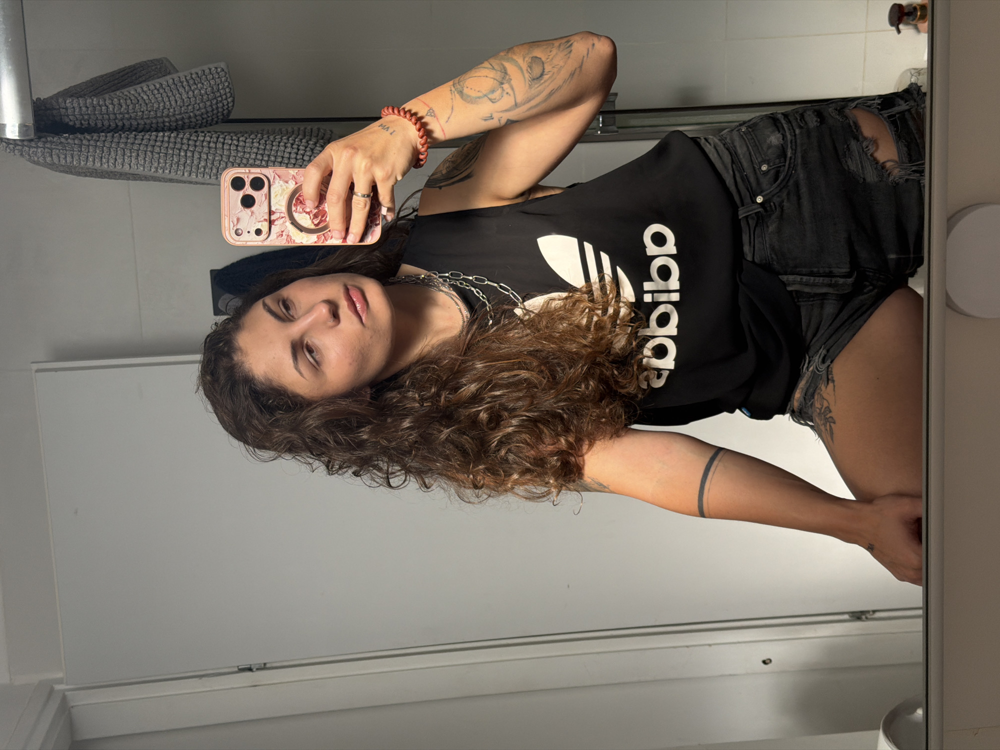 A woman stands in front of a bathroom mirror, taking a selfie. She is wearing a black Adidas t-shirt, ripped black denim shorts, and silver necklaces. She has curly brown hair, tattoos on her arms, and is holding a pink floral-patterned phone. The bathroom has a white door, a towel rack with gray towels, and a white vanity. The location is Kiryat Bialik, Israel.