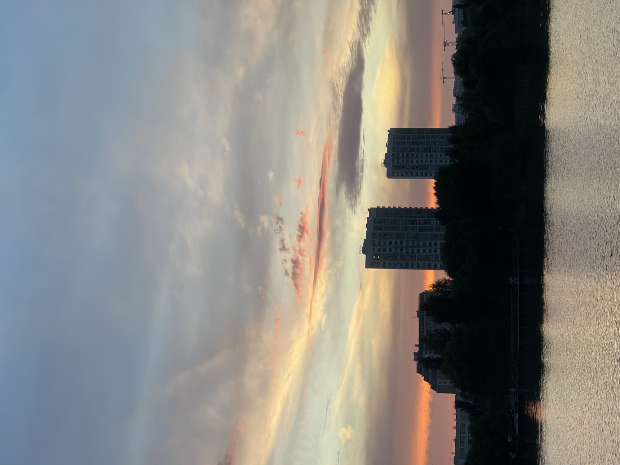 The image depicts a serene lakefront scene at dusk in Moscow, Russia, likely in a residential district such as Maryino, known for its waterfront parks and high-rise buildings. The sky is a canvas of soft pastel colors, with streaks of pink, orange, and pale blue indicating a sunset. Wispy clouds drift across the horizon, catching the fading light.
In the mid-ground, silhouetted against the vibrant sky, are several tall residential buildings. Two prominent, modern apartment blocks stand out, their geometric forms stark against the natural backdrop. Further in the distance, the outlines of more buildings and construction cranes are visible, suggesting ongoing development within the city.
A dark, dense line of trees separates the water from the urban structures, creating a natural buffer. Beneath the trees, a wooden pier extends into the lake, and small figures of people can be seen walking and standing on it, their presence subtle and adding a sense of human activity to the tranquil setting.
The foreground is dominated by the calm surface of the lake. The water reflects the muted colors of the sky and the silhouettes of the trees and pier, creating a shimmering, tranquil effect. The overall atmosphere of the image is one of peaceful urban twilight, where nature and city life coexist.