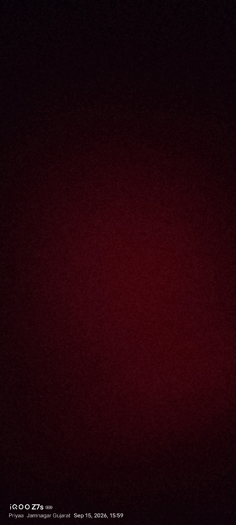 The image is a dark, out-of-focus shot with a gradient of deep red and black. In the bottom left corner, white text displays "iQOO Z7s 5G" followed by "Priyaa Jamnagar Gujarat Sep 15, 2026, 15:59". This indicates the image was taken on September 15, 2026, at 3:59 PM, in Jamnagar, Gujarat, India. The phone model used is an iQOO Z7s 5G. There are no discernible subjects, objects, actions, or interactions visible due to the lack of focus and lighting. The environment appears to be a plain, dark surface, possibly an interior space or a textured background, with no contextual clues about the specific location within Jamnagar.