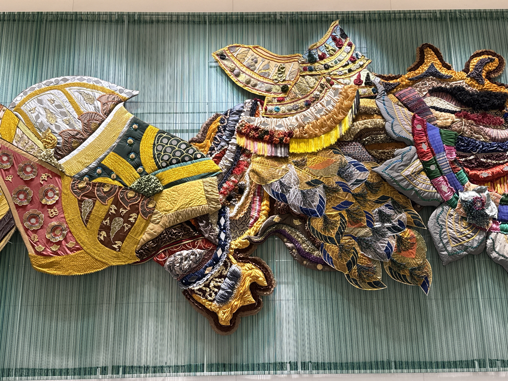 This is a detailed, close-up shot of an elaborate textile art installation displayed against a backdrop of vertical, teal-colored threads or strings. The artwork itself is a richly textured and vibrantly colored assemblage of various fabrics, embroidery, fringe, and embellishments. It appears to be a large, sculptural piece that forms a complex, organic shape, reminiscent of stylized wings or flowing plumage.
The artwork features numerous distinct sections with different patterns and textures. On the left, there are areas with floral motifs in pink and gold, bordered by yellow and green quilted fabric with circular patterns. Moving towards the center and right, the piece becomes more layered and dense. There are sections with intricately embroidered patterns in gold and brown, ruffled fabric in shades of yellow and orange, and woven elements in blues, reds, purples, and blacks. The overall impression is one of opulence and meticulous craftsmanship, with a strong emphasis on three-dimensional elements and a dazzling array of colors and materials.
The artwork is located at the **Siam Paragon shopping mall in Bangkok, Thailand**. Specifically, it is displayed on the wall of the **Art Space, located on the 3rd floor of the S.A.C. (Siam Art & Culture) Gallery at Siam Paragon**. This particular piece is part of the exhibition "Threads of Time" by artist Somruethai Keeratiyut.
