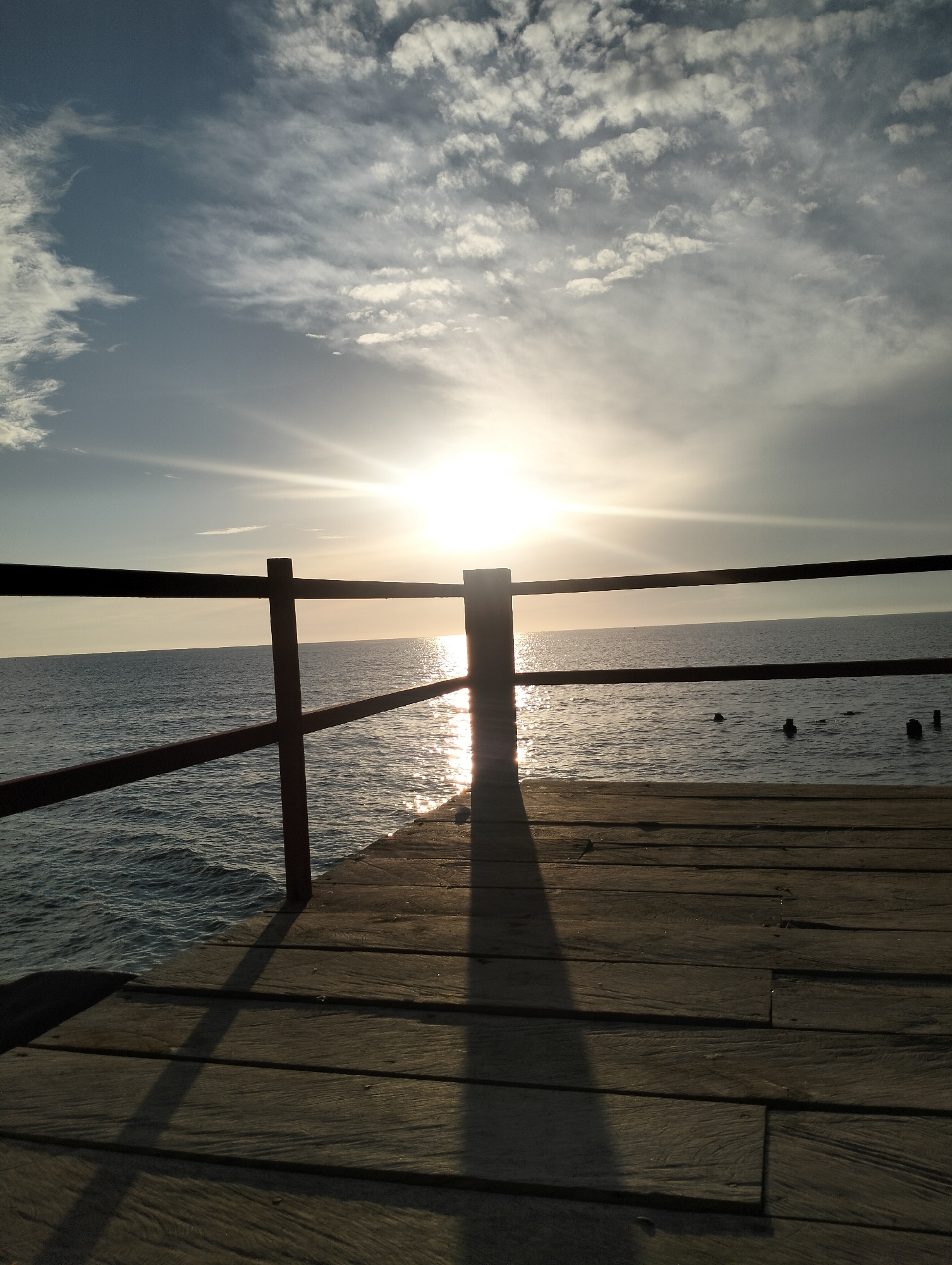 The image depicts a wooden pier extending over a calm, dark blue sea. The sun is low in the sky, casting a bright, reflective glow on the water's surface. The sky is a gradient of pale blue to grey, with wispy white clouds scattered across it. Two dark, vertical posts with horizontal railings are visible in the foreground, partially obscuring the view of the sea and sky. The wooden planks of the pier are weathered and run horizontally across the frame, with a long shadow stretching from one of the posts. A few dark, submerged posts or buoys are visible in the distance on the water. This scene is likely from the coastal area of Lhokseumawe, Indonesia, a city known for its beaches and proximity to the sea. There are no people or animals visible in the image.