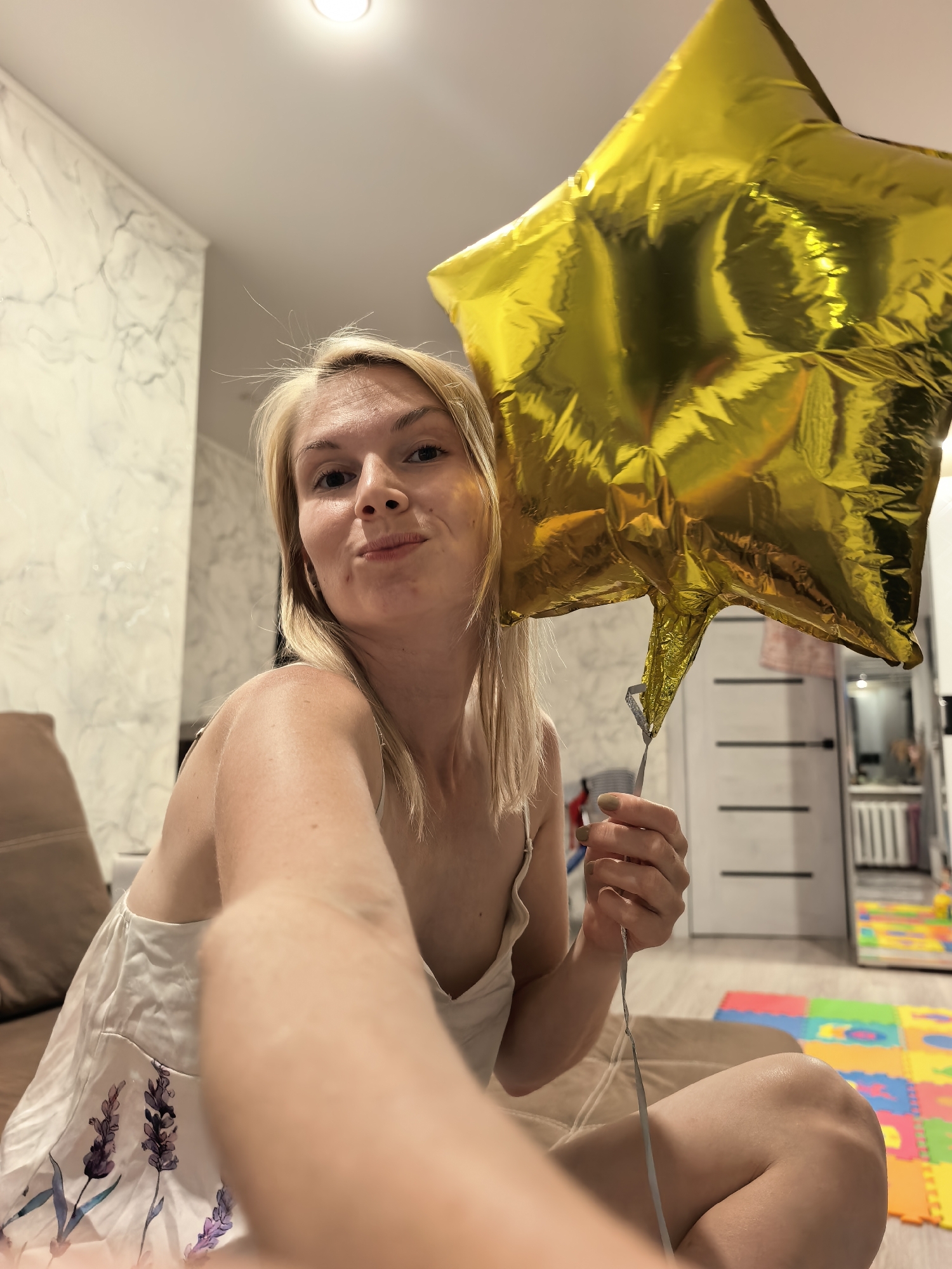 A blonde woman is holding a gold star-shaped balloon indoors. She is wearing a white camisole with a lavender floral print. She is looking at the camera with a playful expression. The background shows a room with marble-patterned wallpaper, a couch, a door, and colorful floor mats. The lighting suggests it is nighttime or indoors with artificial light. There is no information about the location in Sterlitamak, Russia.
