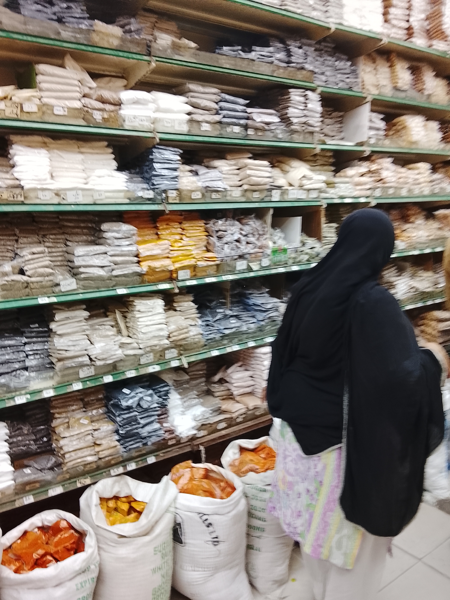 A person wearing a black hijab and a floral pattern skirt stands in front of shelves stocked with bags and containers of various dry goods. The bags on the lower level are labeled "SUGAR RAW WHITE GROSS." The location appears to be a store, possibly in Gulgasht Colony, Pakistan, selling bulk ingredients or spices. The scene conveys a sense of everyday commerce and activity.