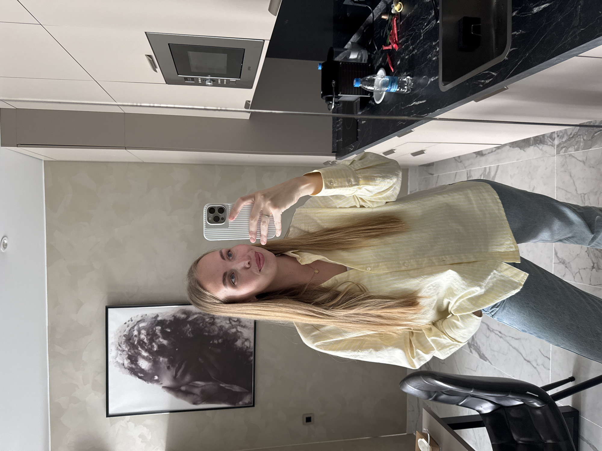 A young woman, likely in her 20s, with long straight blonde hair and light eyes, is captured in a mirror selfie within what appears to be a modern kitchen or apartment in Sochi, Russia. She holds a silver smartphone with a CASETiFY brand ribbed case, extending her arm to take the photo. She is wearing a light yellow and white striped button-up shirt and light blue jeans. A ring is visible on her left hand's ring finger.
The background, visible in the mirror's reflection, features a contemporary interior design. To her left, a framed black and white abstract or textural artwork hangs on a light gray, subtly patterned wall. Above her, built-in light-colored cabinets house an oven or microwave with a metallic handle and a control panel featuring indistinct symbols. A sleek black marble countertop stretches across the back, holding a coffee machine, a partially visible "Evian" water bottle, and some red objects. The flooring consists of light-toned tiles with a marble-like pattern. A black, modern-style bar stool or chair is also visible in the lower right foreground. The overall ambiance suggests daytime, given the soft, even lighting throughout the clean and organized space. The woman has a calm expression with a slight, pleasant smile.