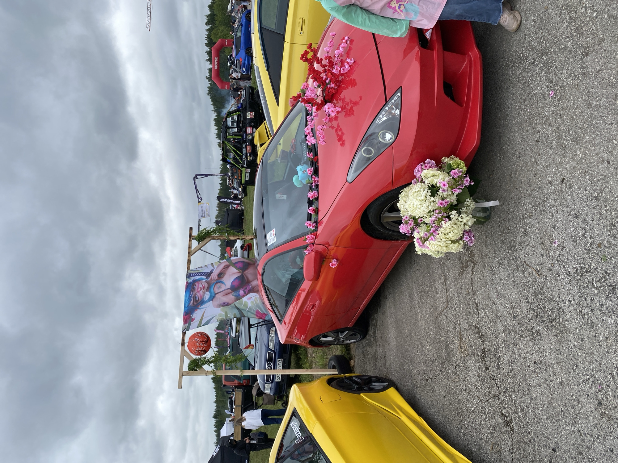 This is a medium shot photograph taken outdoors on an overcast day. The foreground features the front of a red Toyota Celica, which is adorned with pink and red artificial flowers on its hood and dashboard. To the left of the red car is the side of a yellow Toyota Celica. In the background, there are various vehicles, including a dark blue Audi sedan and a green off-road vehicle. A large banner depicting an anime-style character is hanging from a wooden frame. Further in the background, a red inflatable archway with "MOTUL" written on it is visible. The ground is paved with asphalt. The setting appears to be an outdoor event or car show. While the image was taken in the city of Perm, Russia, this information is not visually discernible from the image itself.