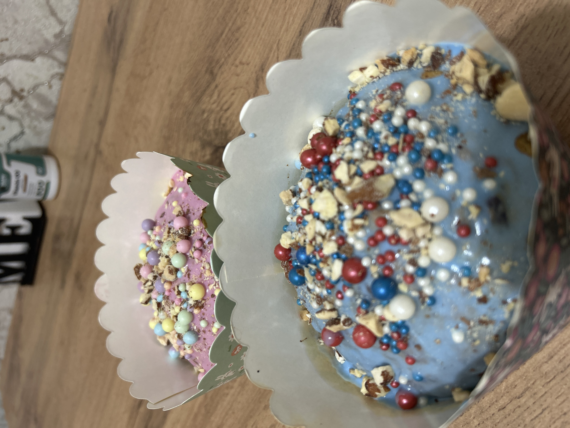 Two cupcakes are displayed on a wooden surface. One cupcake has pink frosting with pastel colored sprinkles, while the other has blue frosting with red, white, and blue sprinkles. Both cupcakes are contained in decorative paper liners. A blurry object is visible in the background to the left of the cupcakes. The scene appears to be indoors. There is no evidence of this image's location within the city of Chaikovsky, Russia.