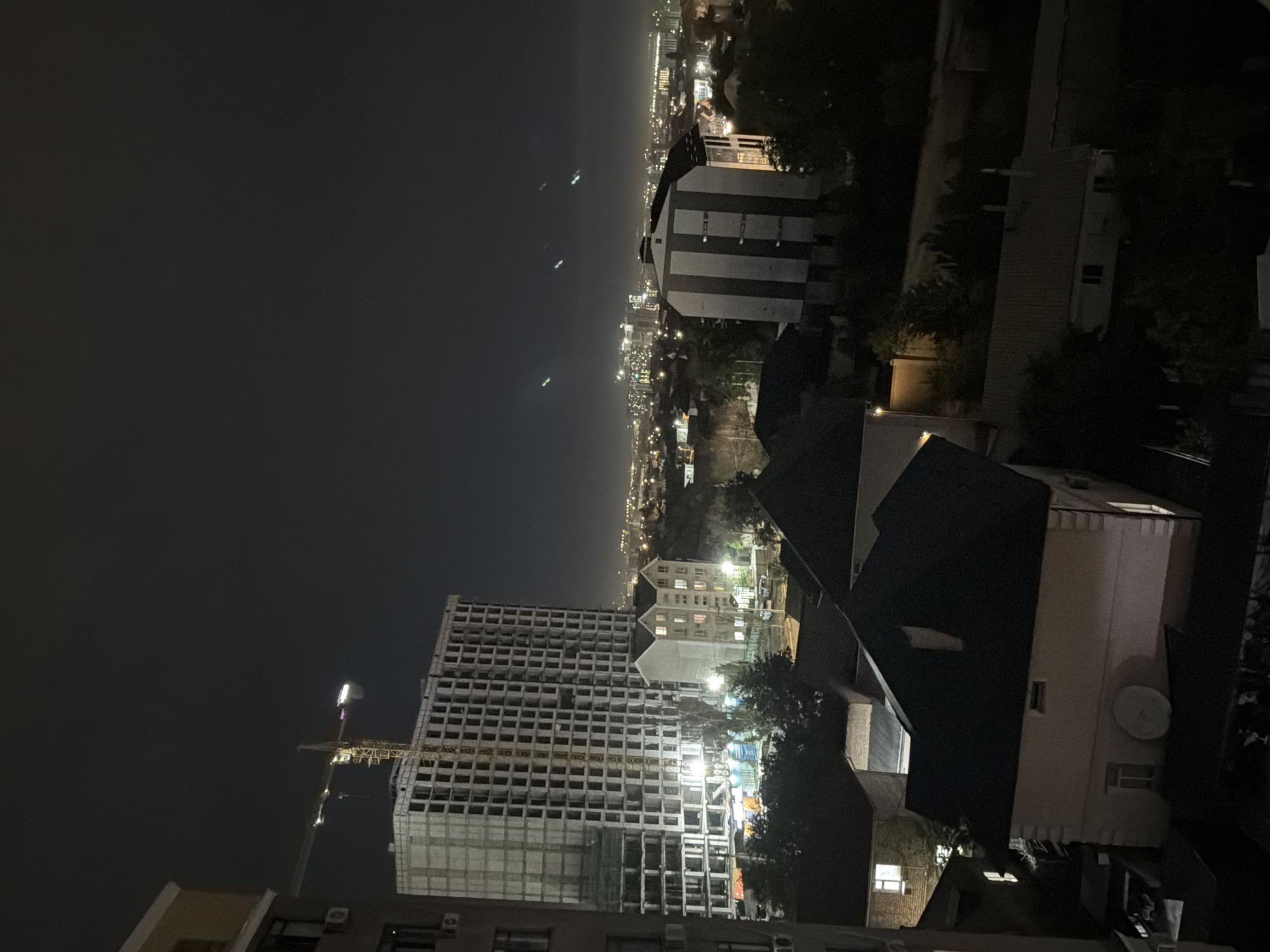 This is a vertical, nighttime aerial shot of a city landscape in Bishkek, Kyrgyzstan. The scene is dominated by buildings, with a large, unfinished high-rise structure in the foreground. This building appears to be under construction, with a prominent crane attached to it. Smaller residential buildings, some with illuminated windows, are scattered throughout the image, creating a mosaic of artificial lights against the dark sky. In the distance, a sprawling urban area is visible, marked by a dense concentration of lights that suggest a larger city center. The sky is dark and overcast, with no stars or moon visible, indicating a cloudy or hazy night. There are faint lights in the distance over what appears to be a body of water, possibly a lake or sea. The overall mood is one of urban development and nocturnal activity, with the illuminated buildings standing out against the darkness. No people are clearly visible, and no text is present in the image.