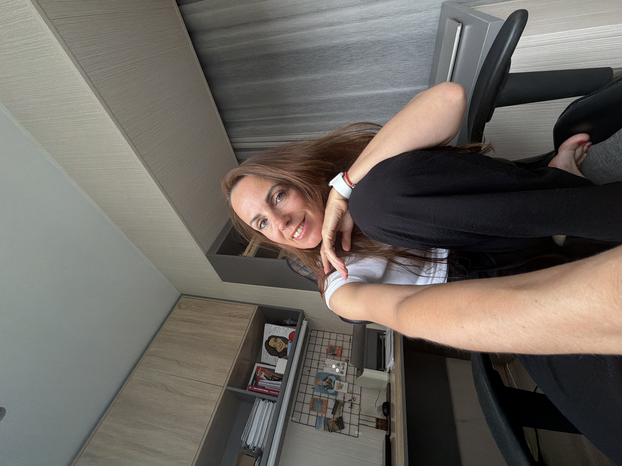A woman is captured in a selfie, holding the camera with her right arm extended. She is seated in an office chair, with her legs crossed. She has long, brown hair and is smiling at the camera, her left hand resting on her cheek. She is wearing a white t-shirt and black pants. A white smartwatch is visible on her left wrist.
The background shows an indoor setting, likely an office or study. There are shelves filled with books and decorative items, including a framed portrait. A wire grid is mounted on the wall, adorned with various pictures and objects. The lighting suggests it is daytime. The overall atmosphere is casual and relaxed.
The original image is rotated 90 degrees counter-clockwise.