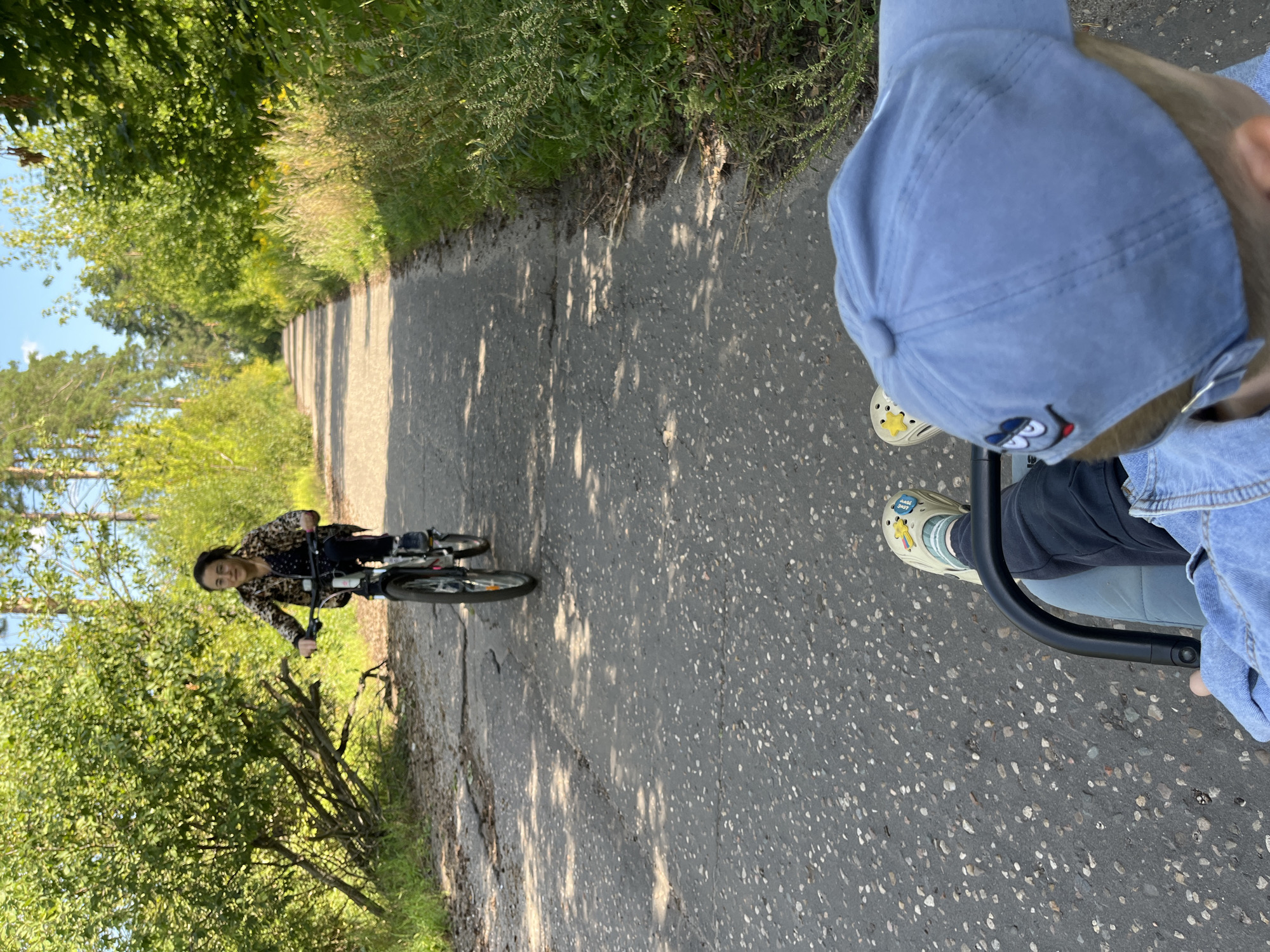 The image depicts a paved path in a wooded area, likely a park or forest trail, in Balashikha, Russia. A woman is riding a bicycle in the background, wearing a patterned shirt and dark pants. In the foreground, a person is seated, facing away from the camera, wearing a blue cap with an embroidered cartoon character, a denim jacket, dark pants, and light-colored crocs with a yellow star charm. The path is cracked and uneven, with grass and trees lining both sides. Sunlight filters through the trees, casting shadows on the path. The overall scene suggests a recreational outing in a natural environment.