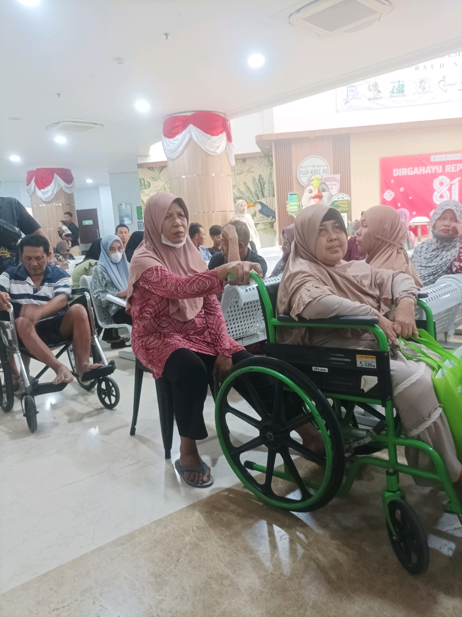 In a brightly lit indoor setting, possibly a hospital or community center in Sumuragung, Indonesia, a group of people are gathered. The floor is a light-colored marble with a reflective sheen. In the foreground, a woman wearing a light brown hijab and a matching kaftan is seated in a green wheelchair. She is looking to her left and appears to be speaking or listening to someone. Another woman in the background, also wearing a light brown hijab and a patterned pink and white top, is seated in a chair and facing to her right. A man in a blue and white striped shirt and dark shorts is seated in a wheelchair to the left of the frame, facing forward. He has dark hair and is looking down. Further in the background, other individuals are visible, some seated and some standing. Decorative red and white fabric drapes are visible hanging from columns in the background, suggesting a festive or formal occasion. A large banner with text in red and white is also present in the background. The overall atmosphere appears calm and orderly.