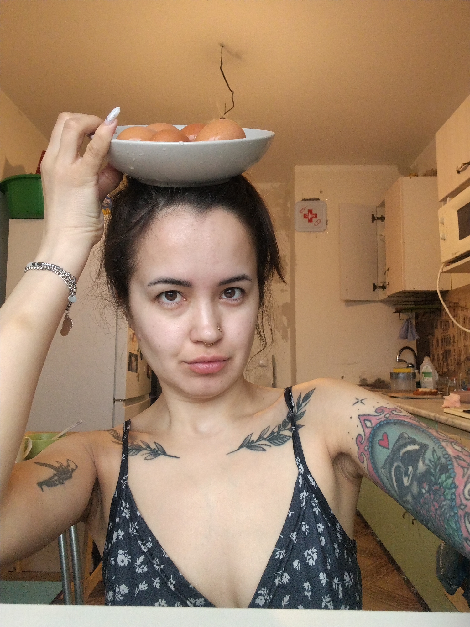 A woman is indoors, possibly in a kitchen in Noviy, Russia. She is holding a white bowl full of eggs on her head. She has medium-length dark hair pulled back and is wearing a patterned sleeveless top. She has multiple tattoos on both arms and chest. The kitchen has white cabinets, a microwave, and a first aid kit on the wall. There is a green bin and a white refrigerator to the left. The countertop is visible on the right with various bottles and objects.