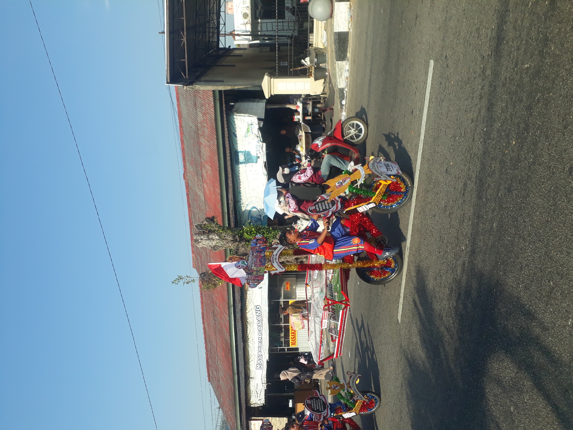 On a sunny day in Pangkalpinang, Indonesia, a street scene unfolds with a parade or festival. The foreground is dominated by a decorated, three-wheeled vehicle that appears to be a modified bicycle or tricycle, adorned with red and gold streamers and a miniature football field display. A young boy in a red and blue soccer-themed uniform rides this vehicle. To the left, another similar vehicle is visible, also decorated and carrying a young boy in a soccer uniform. Behind these vehicles, a red scooter carries two people, one holding a blue umbrella for shade. In the background, buildings with red corrugated metal roofs line the street. A sign above one of the establishments reads "MASAKAN PADANG." People are seen walking and gathered in front of the shops. The sky is a clear, bright blue with a single dark power line stretching across it. The asphalt road shows tire tracks and a white line marking the lane. Shadows from the vehicles and trees are cast across the road.