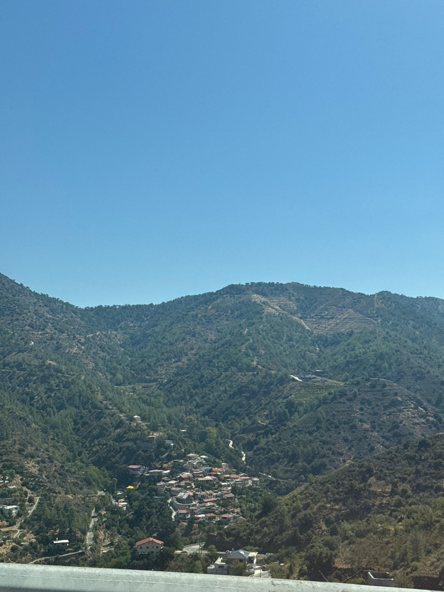 The image shows a panoramic view of Kalopanagiotis, Cyprus, a village nestled in a mountainous region. The sky is a clear, cloudless blue. The foreground is partially obscured by a grey concrete barrier, possibly a highway overpass or retaining wall.
The midground and background are dominated by the lush green slopes of mountains. Dense vegetation, consisting of trees and shrubs, covers the hillsides, with visible winding roads and pathways cutting through the landscape. Scattered across the slopes are numerous houses with red-tiled roofs, indicative of the village architecture. These houses are clustered together in some areas, forming the main village, while others are more isolated.
In the upper part of the mountains, terraced areas are visible, suggesting agricultural activity or land reclamation. The overall impression is of a picturesque mountain village set against a natural, verdant backdrop under a bright, sunny sky. No people are visible in the image.