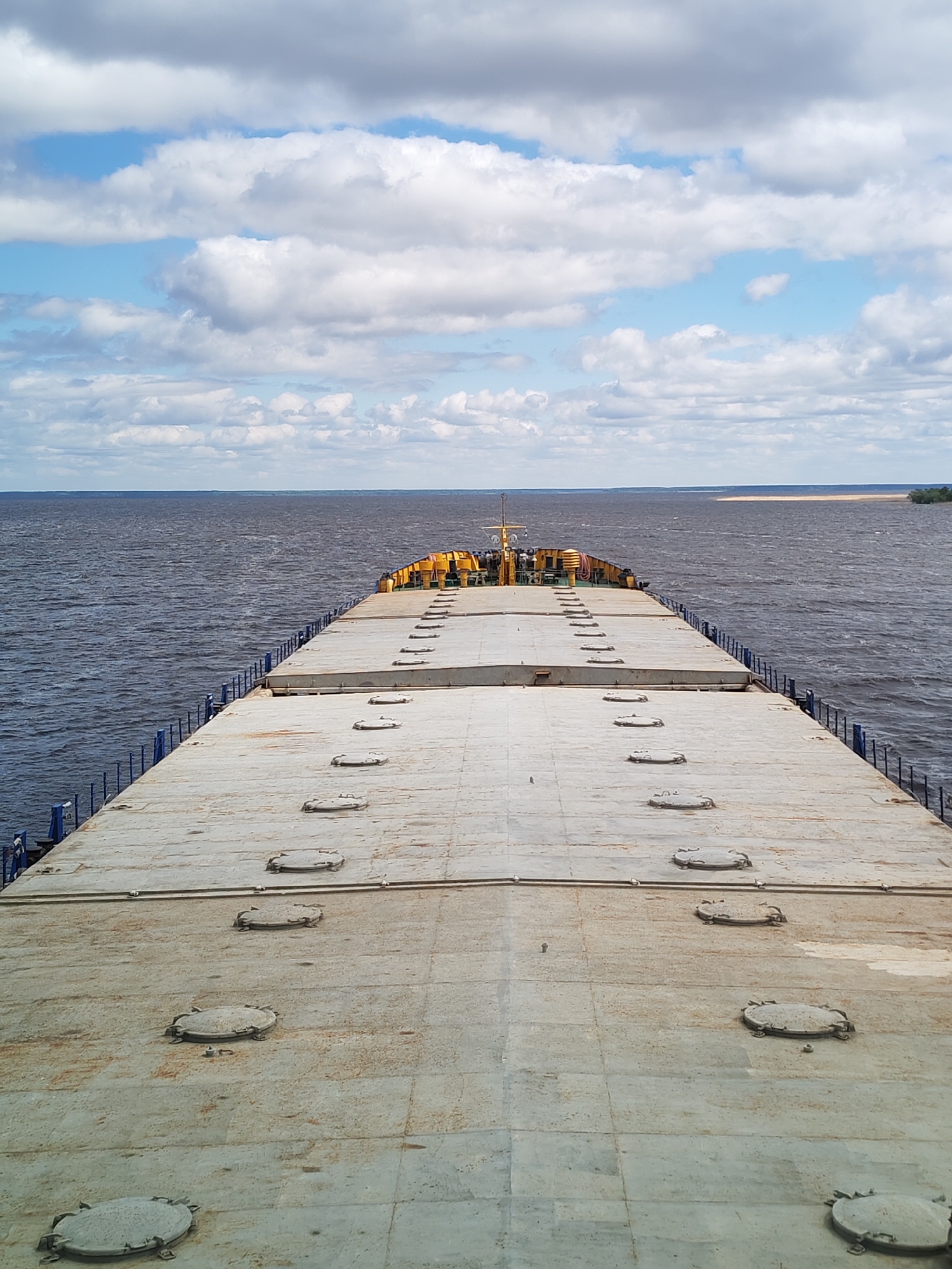 A high-angle shot shows the flat, gray, metal deck of a ship stretching into the distance, with a line of evenly spaced, round access hatches running down the center. The deck is flanked by blue metal railings on either side, beyond which lies a body of water with a muted brown hue. The water extends to the horizon, where land is visible on the right side. The sky is partially overcast with a mix of white and gray clouds, with patches of blue showing through. The location is Unknown, Unknown.