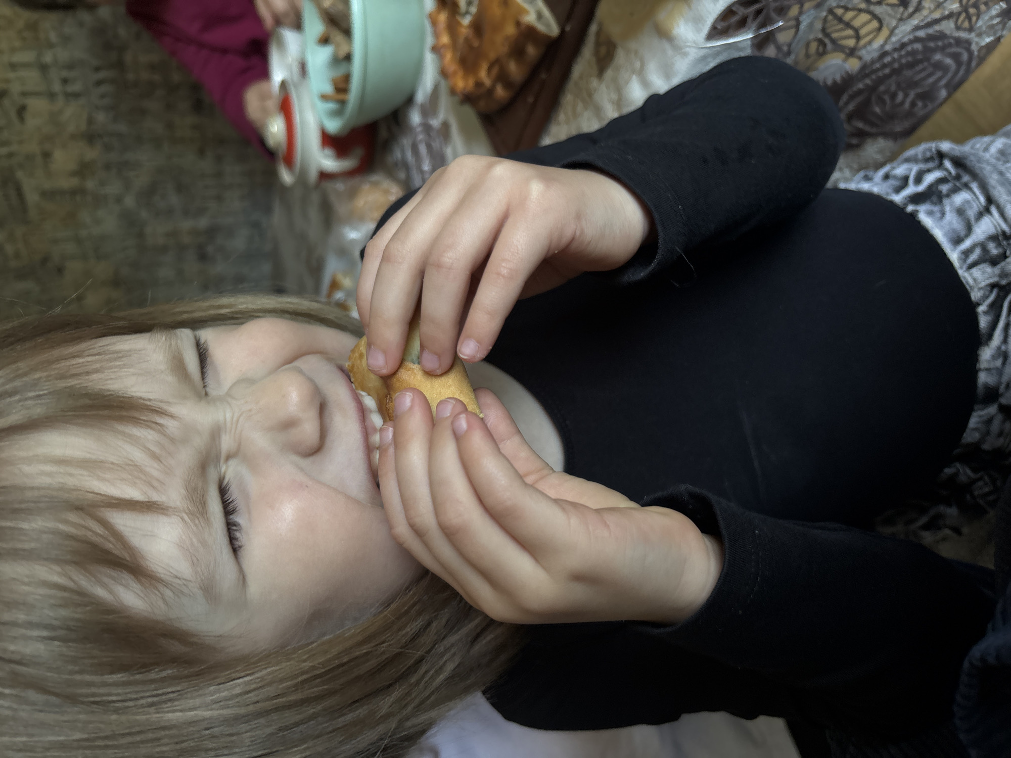 A child with light brown hair is lying down and eating a pastry. The child is wearing a black long-sleeved shirt and grey and white patterned pants. Their eyes are closed and their face is scrunched up as they eat. In the background, there is a light green container with a white lid, a brown pastry on a wooden board, and part of another person wearing a maroon shirt. The location is Аряж, Russia.