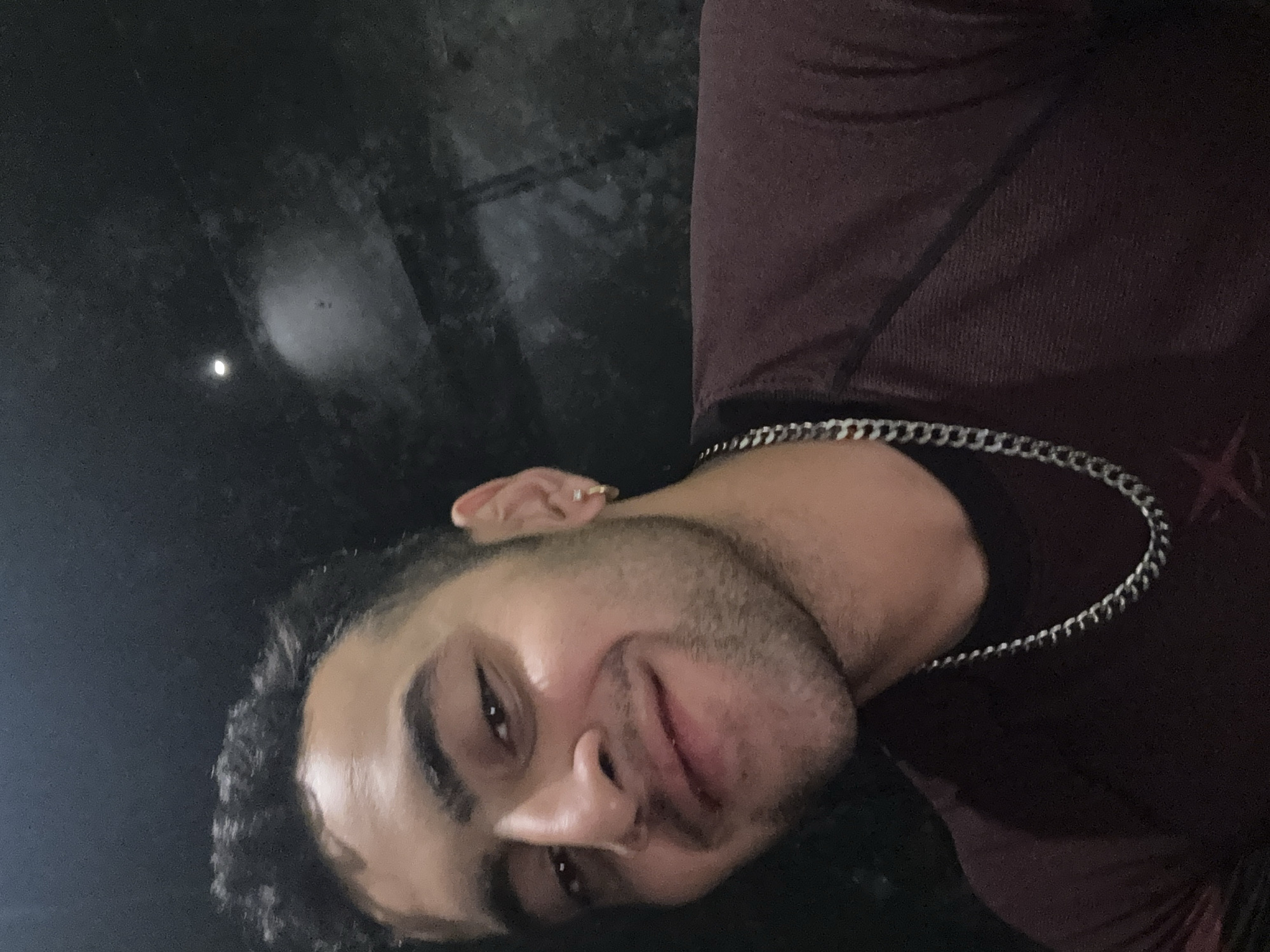 The image displays a head-and-shoulders portrait of a young man, likely a selfie, rotated counter-clockwise. The man has dark, curly hair and a short beard or stubble. He is seen with a subtle, relaxed smile, looking directly towards the viewer. He wears a dark, possibly maroon or burgundy, t-shirt and a silver chain necklace. A small, round, golden stud earring is visible in his right ear.
The background is predominantly dark and appears textured, suggesting a night scene or a dimly lit indoor environment. A distinct, bright, circular light source is visible in the upper left portion of the background, standing out against the darkness. This light could be a distant street light or the moon. The lighting on the man's face is soft and diffused, creating some shadows but clearly illuminating his features.
There are no discernible activities taking place other than the posing for the photograph. No specific location cues are visible within the image itself to confirm a location like Kolkata, India, though the subject's appearance is consistent with the region. No visible text is present in the image. The overall impression is one of a casual, personal moment captured in low light.