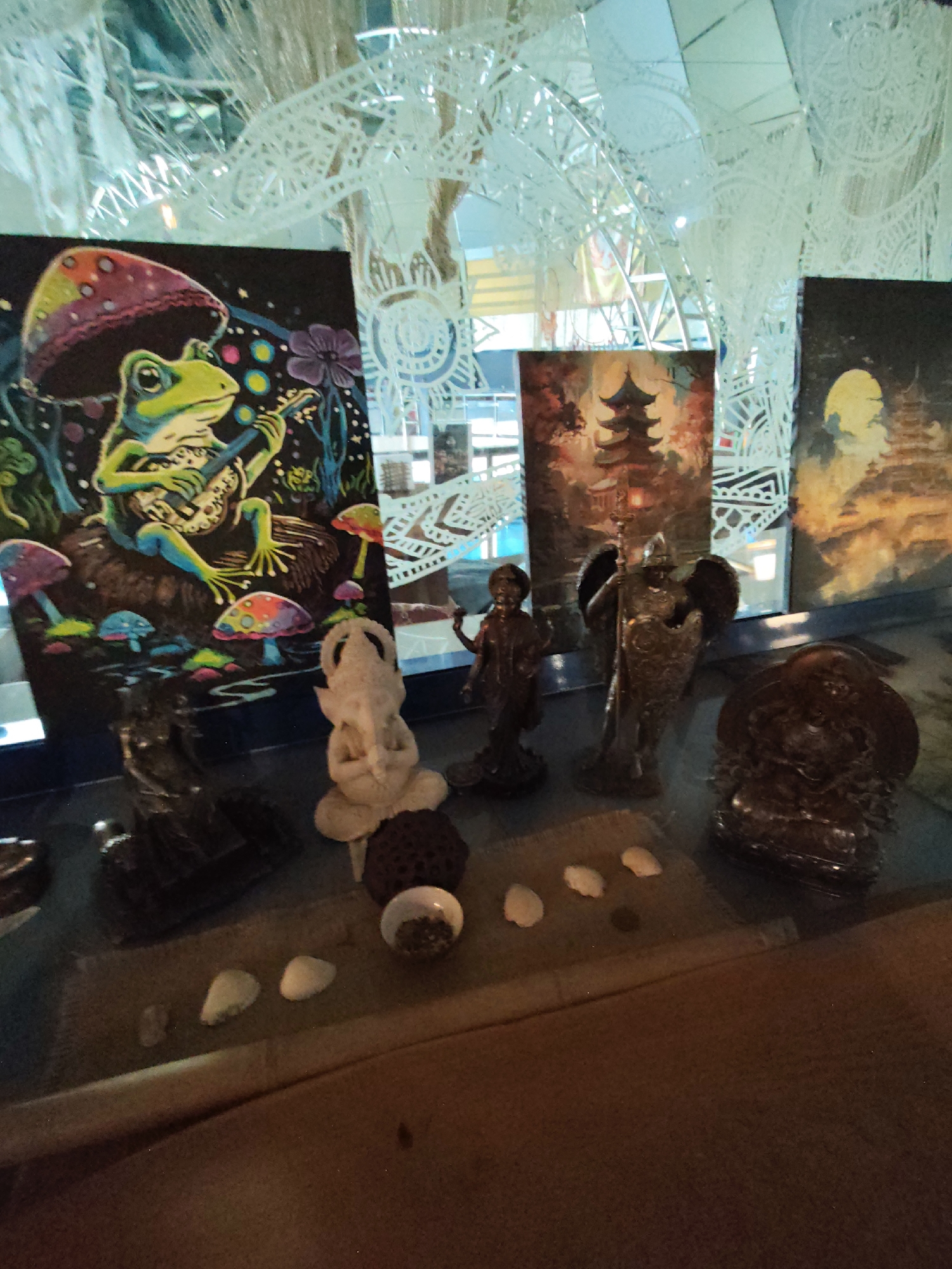 The image depicts a display of artistic items, including paintings and figurines, arranged on a shelf or table in what appears to be an indoor setting. In the background, a decorative, translucent structure with intricate patterns is visible. To the left, a painting features a frog playing a guitar amidst psychedelic mushrooms and flowers. To the right of this, a painting of a pagoda is partially visible, followed by another painting of a pagoda silhouetted against a moonlit sky. In front of the paintings, several figurines are displayed: a light-colored, stylized figurine resembling an elephant or a creature with tusks, a dark bronze-colored figurine of a winged warrior holding a spear and shield, a smaller, dark bronze-colored figurine of a standing figure, and a seated dark bronze-colored figurine with multiple arms. Scattered on the surface in front of the figurines are small, light-colored objects that appear to be shells or stones. The overall scene suggests a collection of decorative and artistic objects, possibly for sale or display in a shop or personal collection. The image does not contain any discernible elements that would place it specifically in the city of Sochi, Russia.