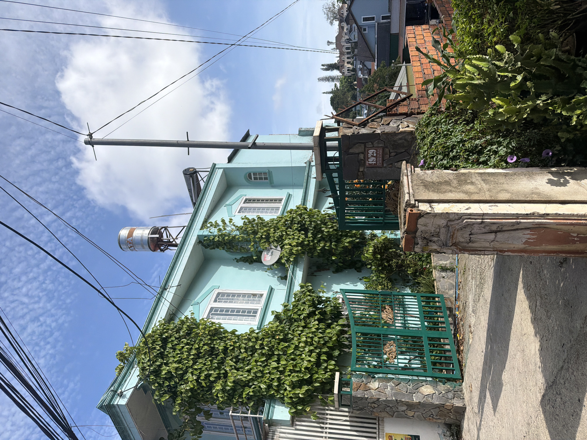 A bright, slightly underexposed, medium-angle outdoor shot captures a turquoise house on a sunny day in Da Lat, Vietnam. The house, adorned with abundant green ivy cascading down its walls, features white-framed windows and a balcony. A metal water tank sits atop the roof, and a solar water heater is visible on the upper level. A dark green metal gate, embellished with decorative dragon motifs, is partially open, revealing a stone-pillared entrance. The gate is flanked by stone-clad pillars and a wall constructed of brick. A sign on one of the pillars reads "49/29". Overhead, a tangle of black electrical wires stretches across the frame against a blue sky dotted with white clouds. The foreground consists of a cracked concrete path, casting shadows. In the background, a cluster of buildings with pitched roofs can be seen nestled amongst the trees, suggesting a hillside urban environment. The overall impression is one of a residential street scene in Da Lat, characterized by its lush vegetation and distinctive architecture.