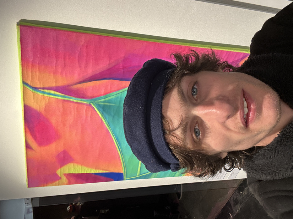 A fair-skinned person wearing a dark blue cap and a black fleece jacket poses in front of a colorful abstract artwork. The artwork features shades of pink, orange, yellow, green, and blue, with a geometric design. The person has light blue eyes, wavy brown hair, and a slight smile. The image is taken at a close-up angle.