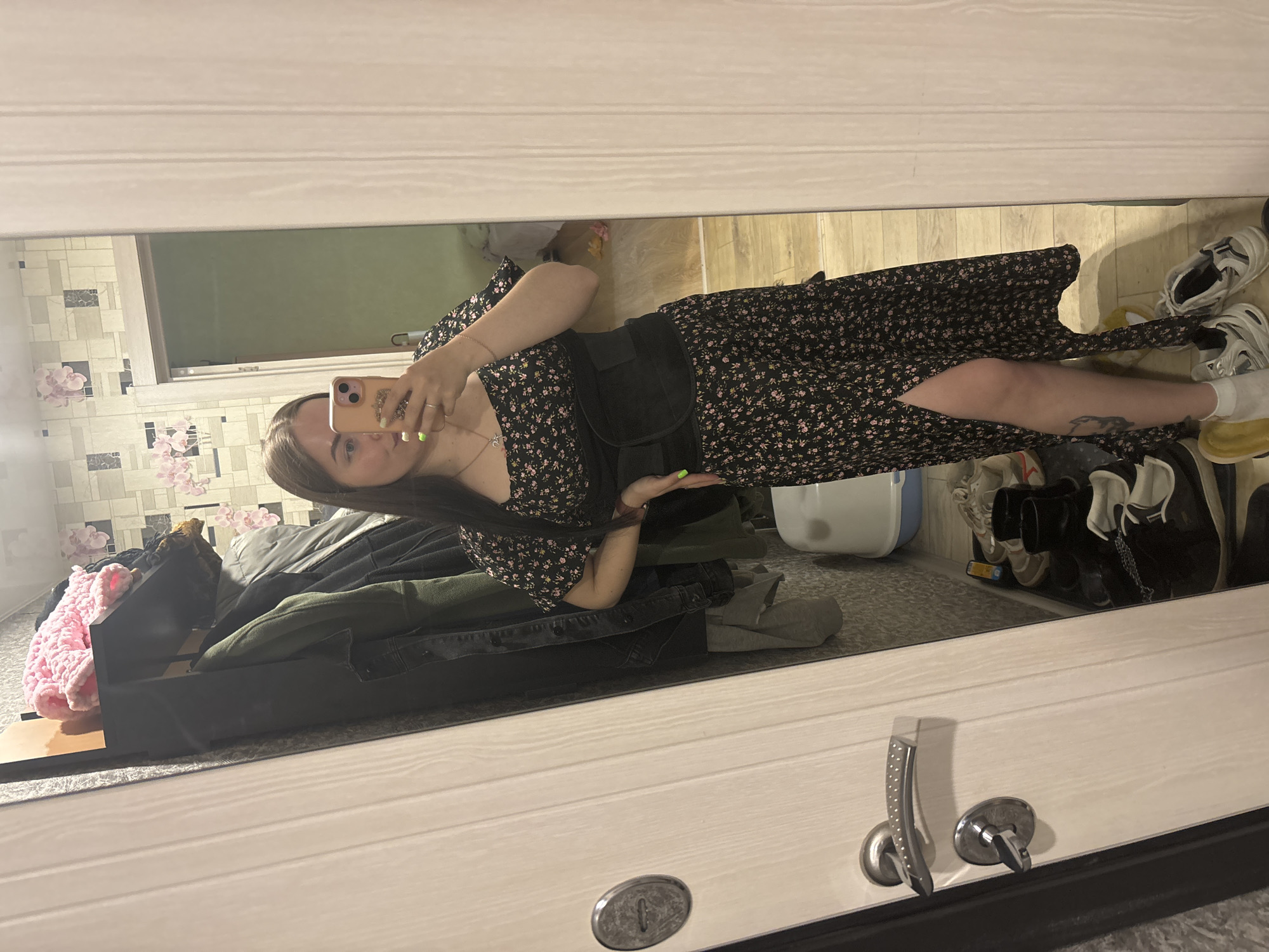 A woman takes a mirror selfie in a room in Izhevsk, Russia. She wears a floral dress, a black waist trainer, and white socks. Her left leg is visible through the dress's slit, and a tattoo is visible on her left calf. She holds her phone up to capture the image. The mirror reflects a room cluttered with clothes, a laundry basket, and shoes. The background wall is decorated with a floral pattern. The mirror itself is attached to a white door with a silver handle and lock.