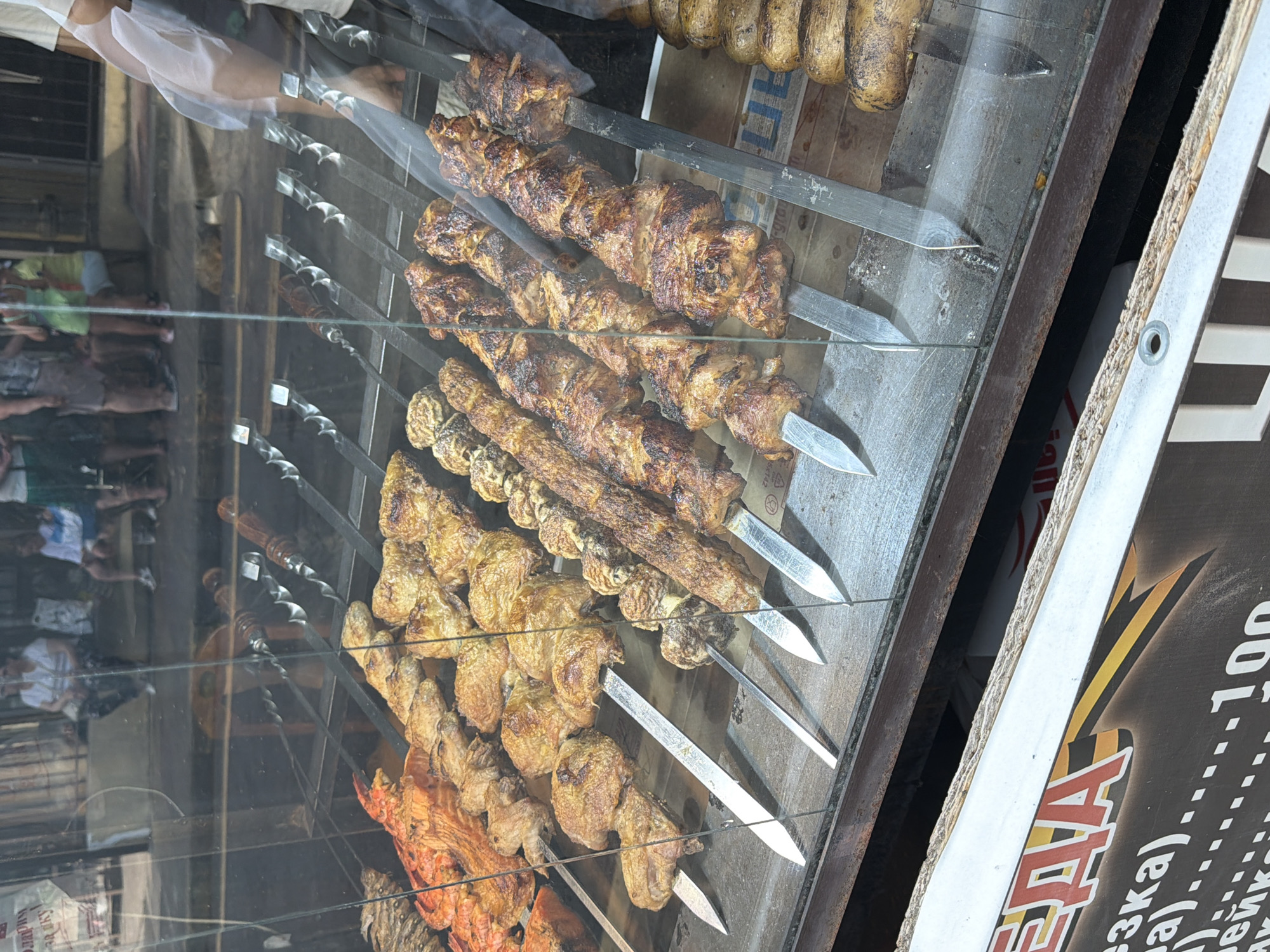 This is a medium-shot, eye-level photograph taken through a glass display case, likely in Sochi, Russia. The foreground features several rows of skewers laden with cooked meat and seafood. The meat appears to be a mix of chicken and possibly lamb or pork, grilled to a golden brown and some pieces showing char marks. The seafood closest to the viewer appears to be a large grilled shrimp or lobster. Behind the meat skewers, more skewers of what looks like meat are visible. To the right, a row of whole roasted potatoes on skewers is present. The display case is made of glass with a wooden trim at the bottom, and a portion of a dark brown wooden shelf is visible below the glass. Reflected in the glass are blurred figures of people, some appearing to be patrons standing or sitting in the background. The overall setting suggests an outdoor food stall or restaurant. The image has a slight glare from the glass, obscuring some details.
