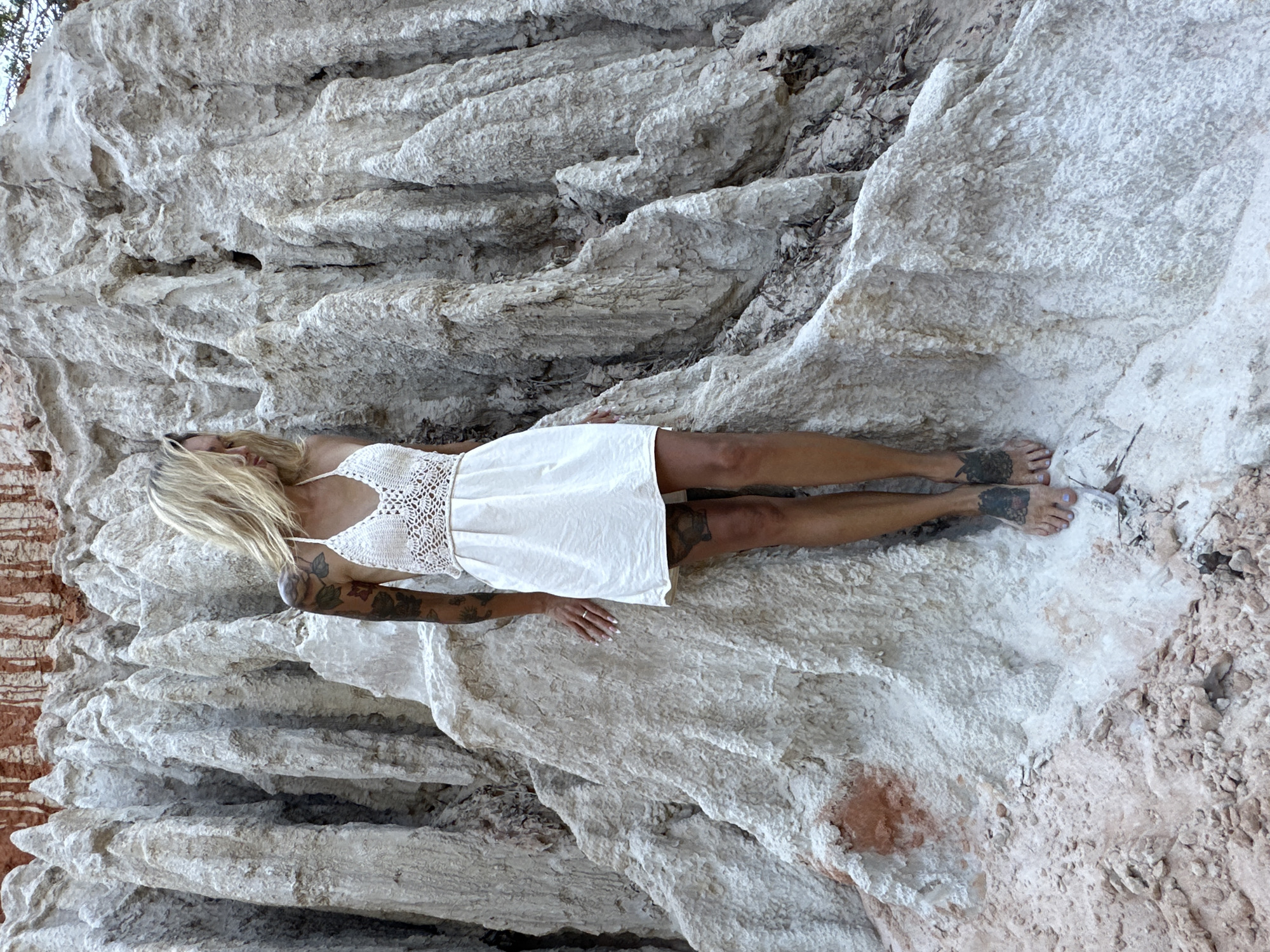 A blonde woman stands on a white, textured geological formation in the Fairy Stream in Phan Thiet, Vietnam. She is wearing a white crochet top and a white linen dress. The woman has tattoos on her arms and legs. She is looking down and to her right. The background consists of the same white geological formations, with some reddish-brown rock visible at the top. The ground is covered in sand and small rocks.