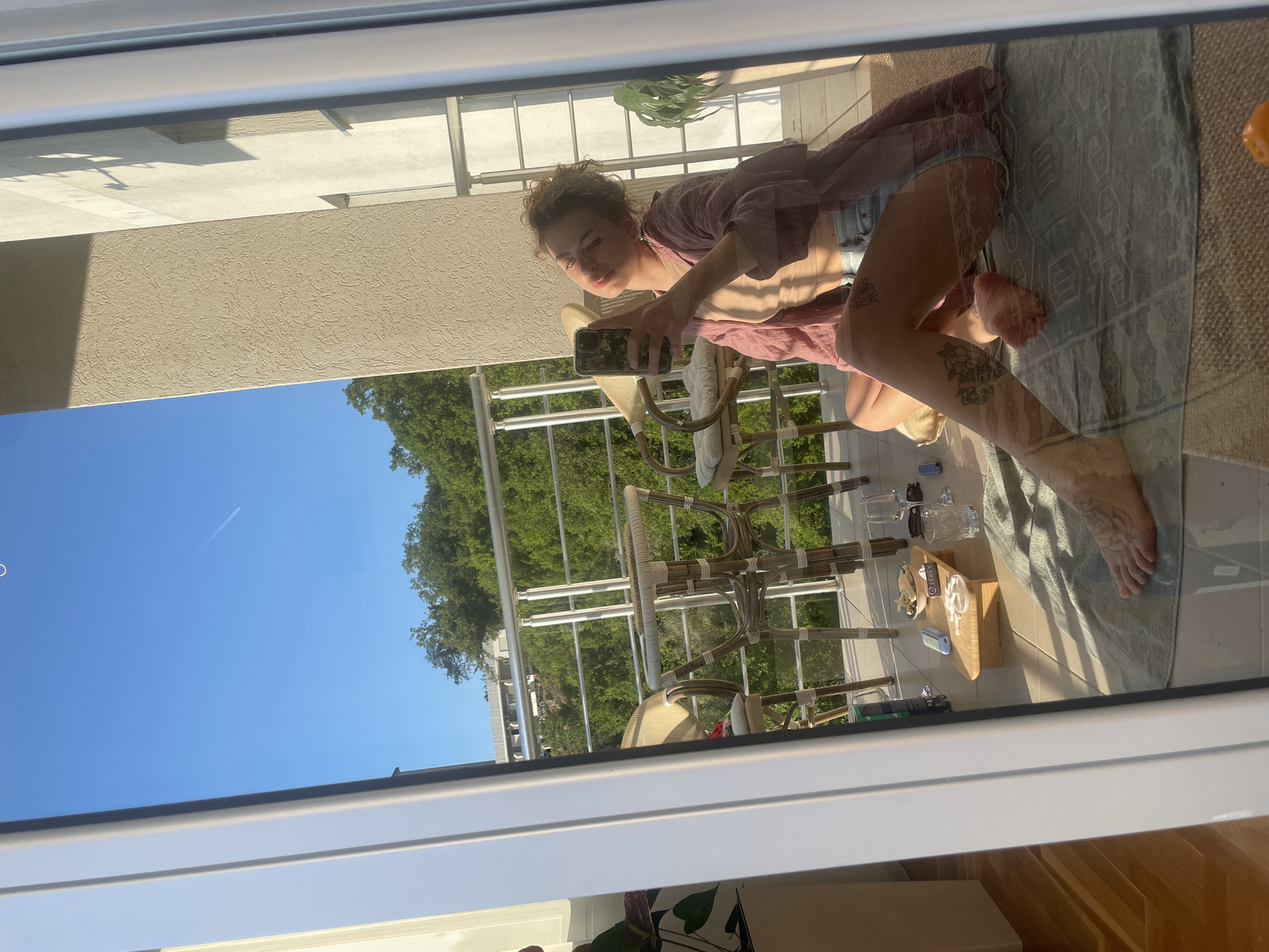 The image shows a reflection in a window of a woman taking a selfie on a balcony. The balcony appears to be in Rafailovići, Montenegro, given the visible buildings and surrounding greenery. The woman is kneeling on a patterned rug, wearing a loose pink shirt and denim shorts. She is holding a smartphone in her hand, presumably taking a picture. Behind her are woven chairs and a small wooden table holding glasses, a box, and other items. The reflection also shows trees, a clear blue sky, and portions of adjacent buildings.