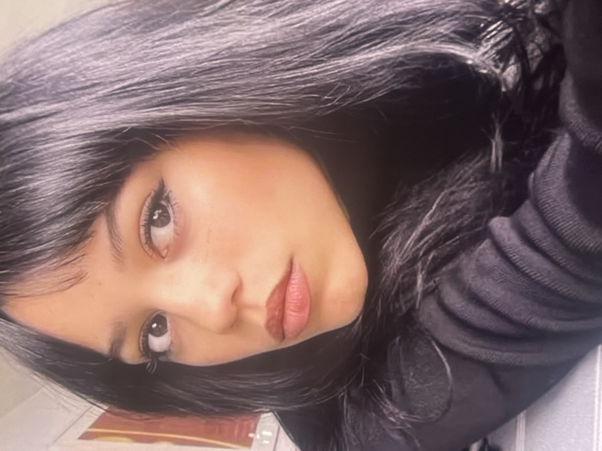 The media file presents a close-up portrait of a young person, likely female, captured in a selfie-style photograph. The image is notably tilted, featuring the subject's head at an angle. She has long, straight, dark hair, possibly black, which cascades around her face and shoulders, with strands forming a fringe across her forehead.
Her facial features are prominent: dark eyes, possibly brown, are adorned with long, dark eyelashes, suggesting the use of mascara or false lashes. Her eyebrows are dark and well-defined. Her nose is straight, and her full lips are painted with a muted reddish-brown or berry-toned lipstick, held slightly parted. Her complexion appears smooth, with a hint of blush on her cheeks.
The subject is wearing dark clothing, likely a sweater or hoodie, visible around her shoulders and neck, featuring a ribbed or textured fabric. The background is predominantly dark and out of focus, providing minimal environmental details.
The person's expression is neutral, appearing contemplative or subtly posed for the camera. There are no explicit visual cues within the image to indicate the specified location of Poltava, Ukraine. The lighting suggests an indoor setting, with soft illumination on the subject's face. No specific activities are depicted other than posing for the photograph, and no text is visible in the image.