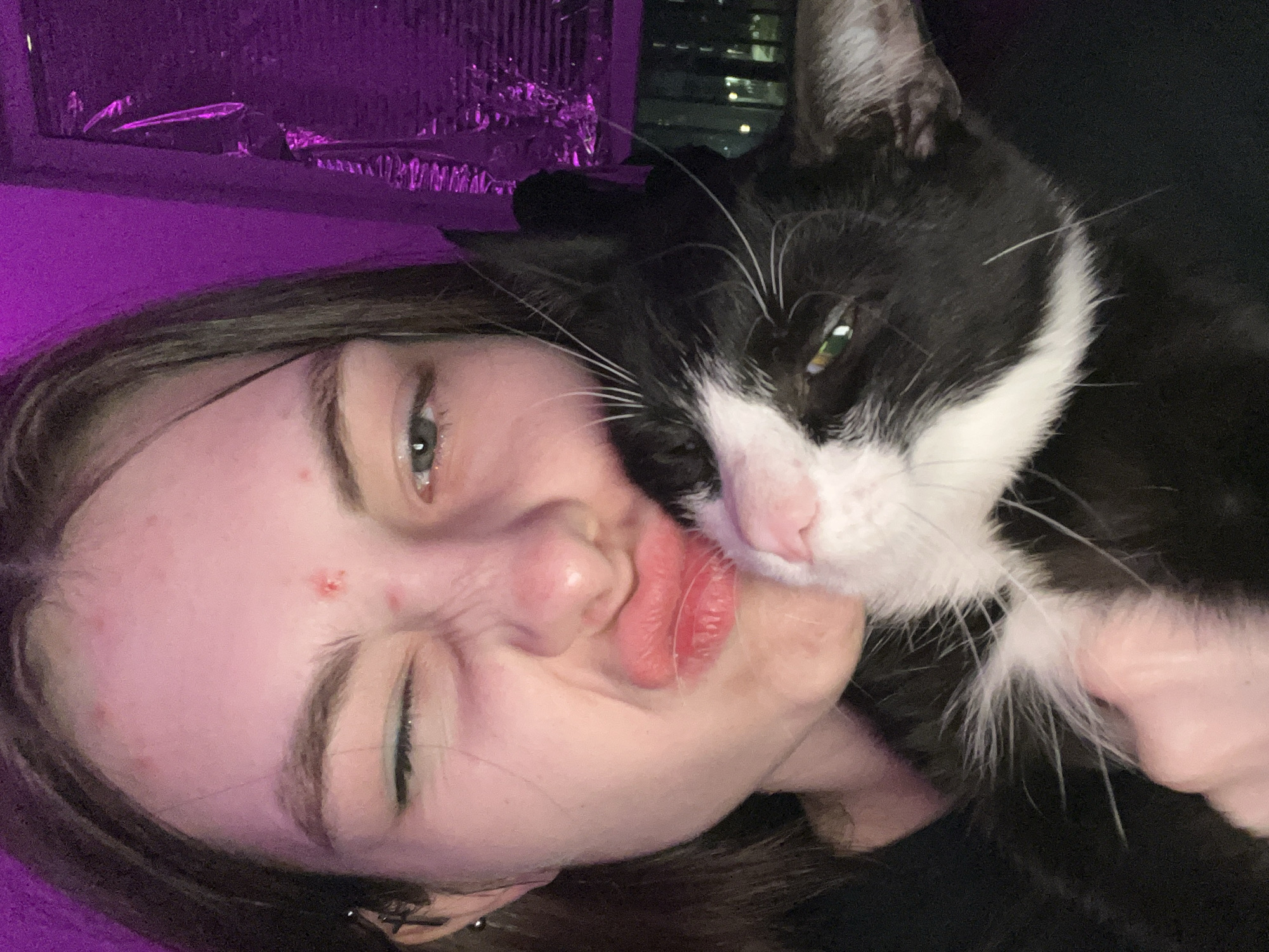 A young woman is holding a black and white cat to her face. She is pouting her lips and squinting one eye. The background is dark, with some light visible from a window. The scene does not depict any details specific to Saint Petersburg, Russia. The information about the location cannot be confirmed from the image.