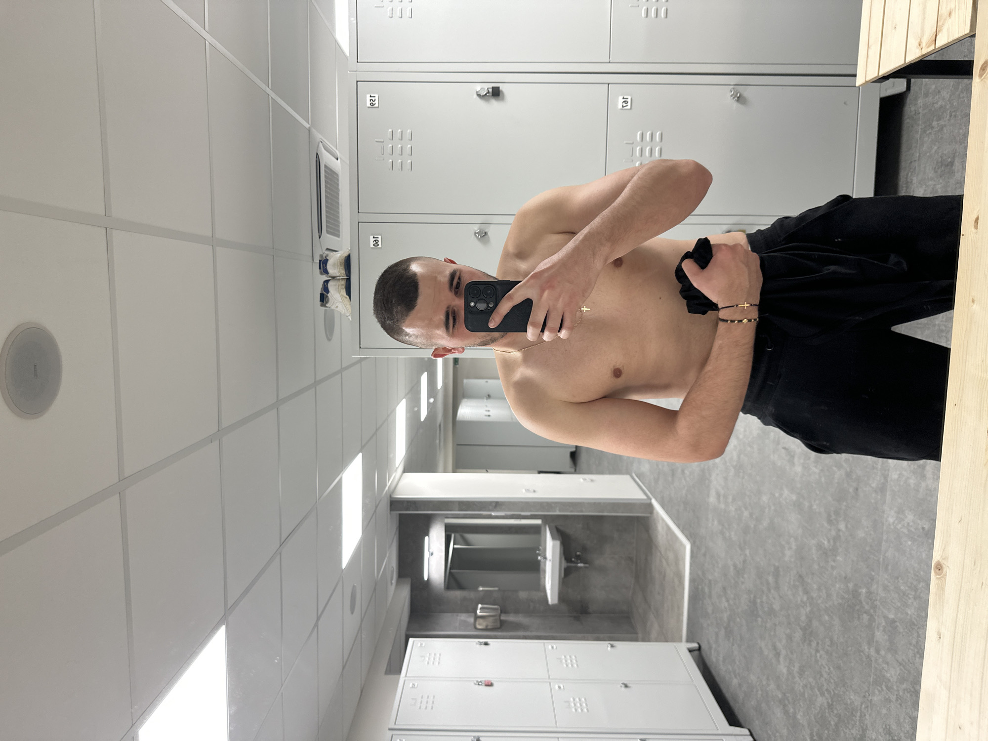 A shirtless man is taking a selfie in what appears to be a locker room. He is wearing black pants. The locker room has gray lockers along one wall, a sink and mirror area in the back, and gray tiled floors. The location cannot be determined to be in the city of Plovdiv, Bulgaria, without additional information.