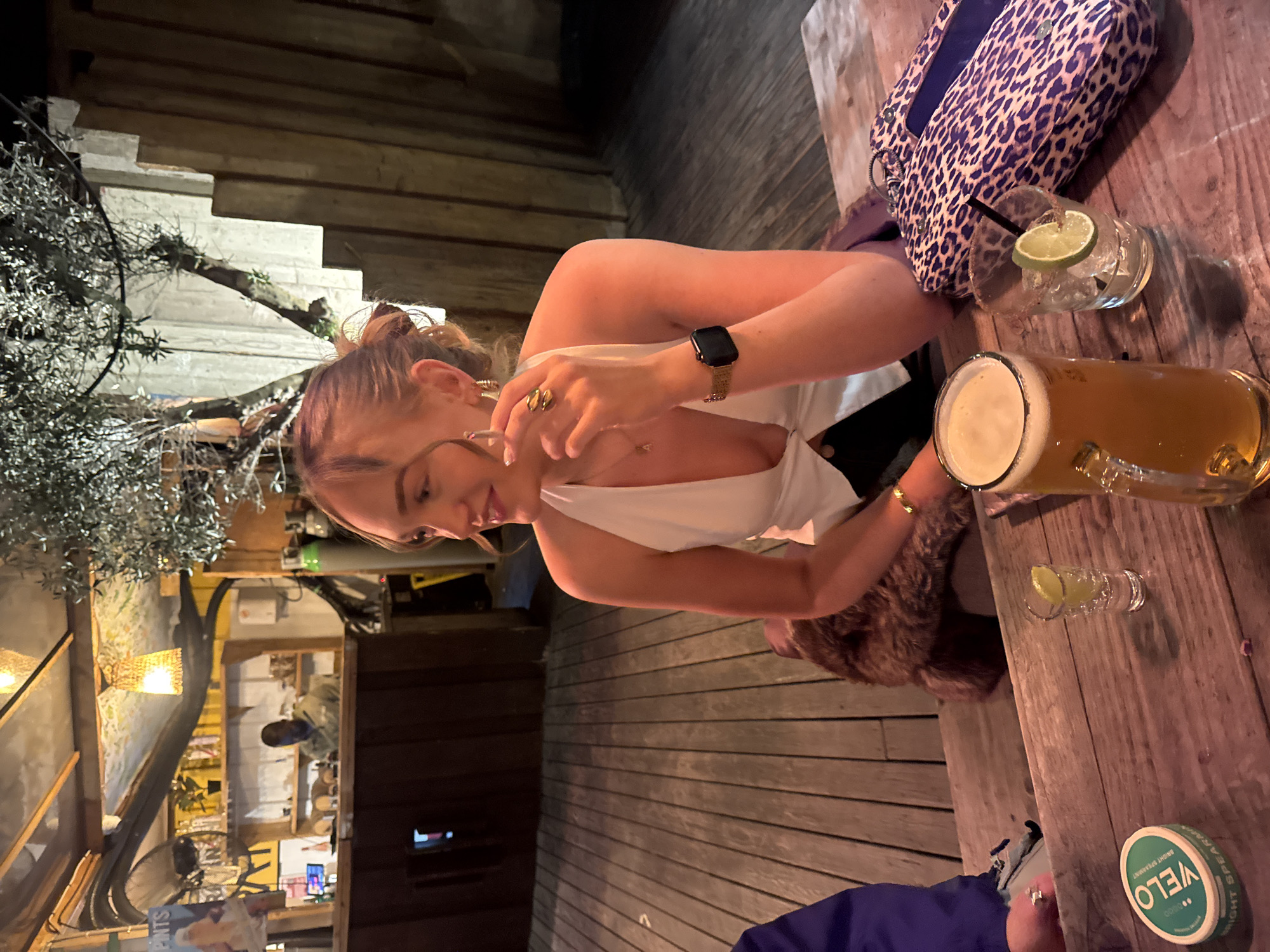 The image captures a casual evening scene, likely at a bar or outdoor patio in London, United Kingdom. Two individuals are seated at a rustic wooden table, enjoying drinks.
The primary subject is a woman with light brown hair, pulled back in a bun, wearing a white V-neck top. She has a watch with a black face and a woven strap on her left wrist, along with several gold rings on her fingers. She is leaning forward, looking down at the table with a slight smile, suggesting a relaxed and pleasant mood.
On the wooden table, there's a large glass of beer with a foamy head and a smaller clear glass containing ice and a slice of lime, possibly a cocktail. A small shot glass with a lime slice is also visible. Next to the drinks, a distinctive leopard print clutch bag rests on the table. Closer to the viewer, a green circular tin with text "VELO" and "BRIGHT SPEARMINT" is present.
The background reveals a warm, dimly lit environment, indicative of evening or night. Wooden planks form the floor or decking, and further back, a light-colored structure that resembles stairs or a tiered wall is visible, entwined with branches from an olive tree. Other background elements include various wooden structures, a wicker light fixture, and glimpses of what appears to be a bar area with shelves and bottles in the distance. Part of a bicycle frame is also faintly visible in the background, along with a partially visible poster that includes the word "PINTS". A second person with brown, curly hair is partially visible next to the main subject.