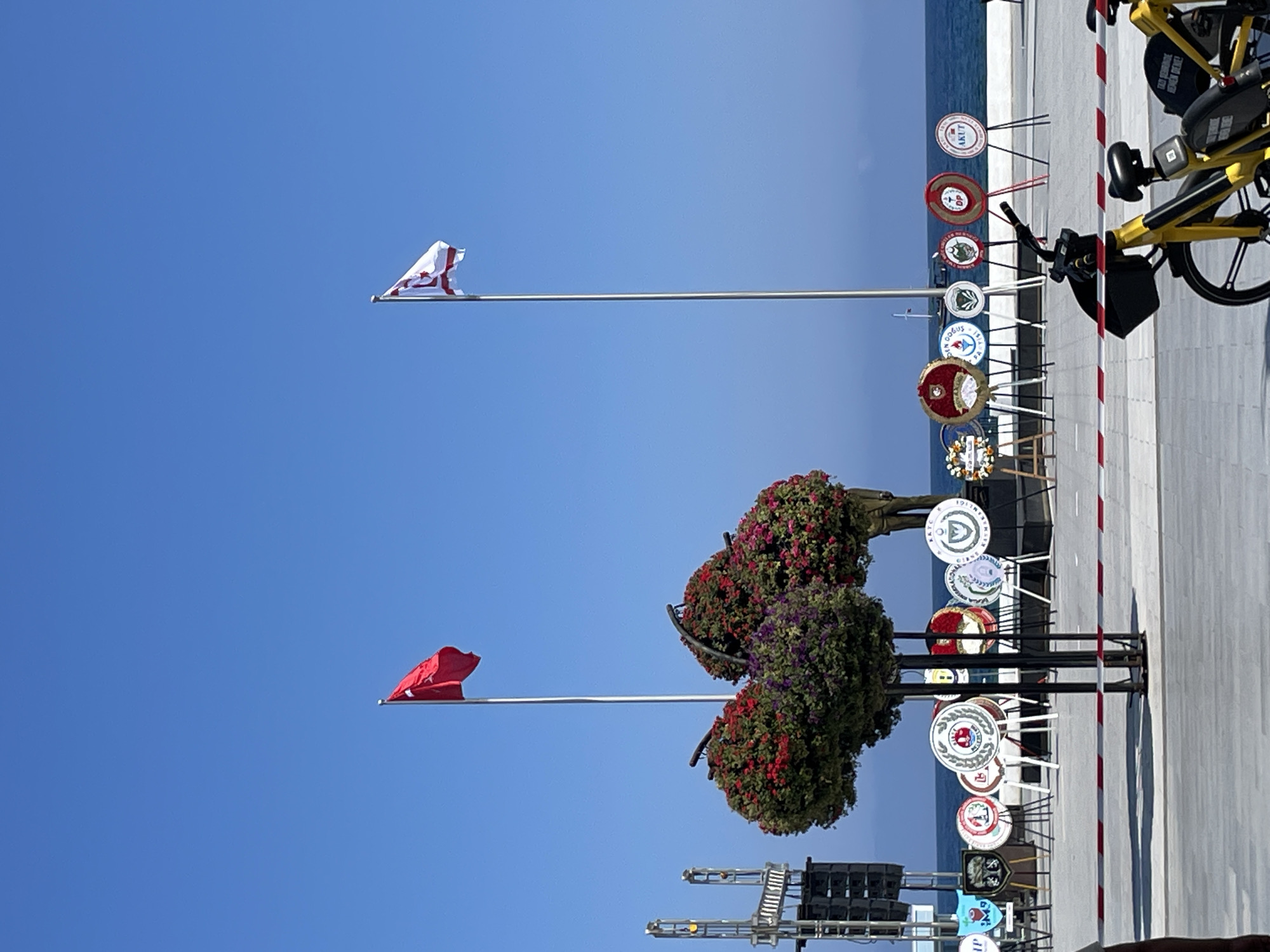 The image shows a public space near the sea in Kyrenia, Northern Cyprus. Two flags are flying on poles; one is red with a white crescent and star, and the other is white with red stripes and a red symbol. In the foreground, a line of circular wreaths on stands is arranged. In the middle ground, there are two elaborately decorated floral displays, one with red and purple flowers. Behind the wreaths, the sea is visible with a boat in the distance. In the far background, there is a clear blue sky. Partially visible in the lower right corner are two yellow bicycles, one with a basket.