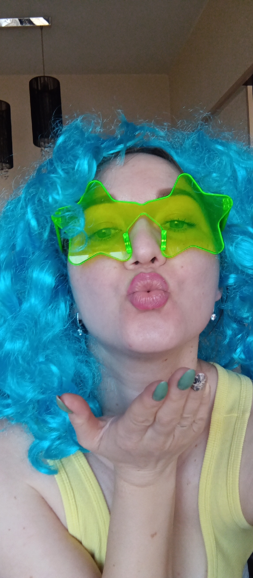 A fair-skinned woman with a blue curly wig and yellow star-shaped glasses puckers her lips in a kiss towards the viewer. She wears a yellow tank top, and her nails are painted green. She has a silver ring on her finger and dangling earrings. The background includes a hanging light fixture. The location is a building.