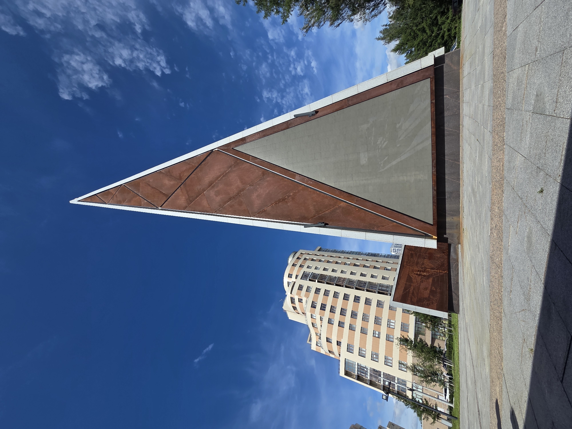 An external, low-angle, wide shot captures a modern, triangular monument in Yekaterinburg, Russia, against a bright blue sky dotted with wispy clouds. The monument features a large, triangular panel, likely a screen or plaque, framed by reddish-brown metal panels with a geometric pattern and white trim. To the left of the monument, a tall, contemporary building with a curved facade and beige and tan brickwork rises prominently. The surrounding ground is paved with large, grey granite tiles, with a decorative band of smaller, reddish-brown tiles in the foreground. Trees, including evergreens, are visible to the right and behind the monument. The scene is devoid of people or any visible activity.