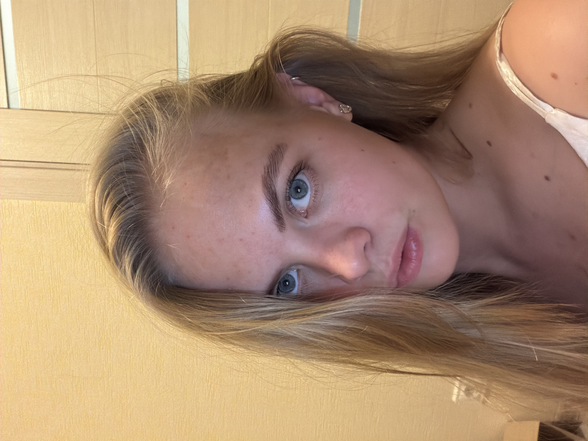 A close-up, tilted portrait photograph depicts a young woman with light skin, blonde hair, and blue eyes. She appears to be wearing a light-colored top. The background consists of a pale yellow wall and a portion of what appears to be a wooden cabinet or door. No specific location in Izhevsk, Russia, is discernible from the visual information provided in the image. The image contains no discernible actions or interactions.