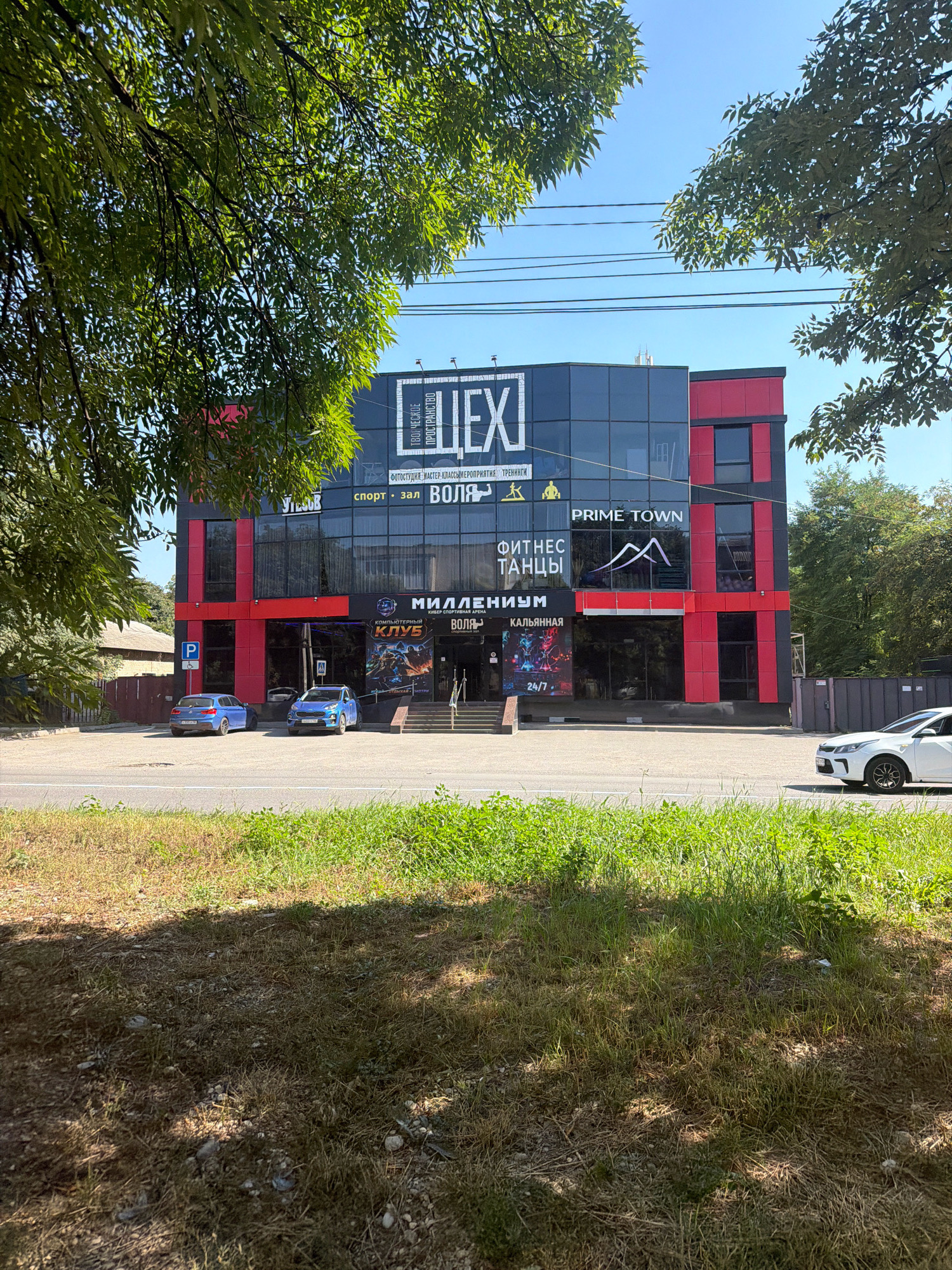 The image captures a modern, multi-story building located in Yessentuki, Russia. The building features a striking facade with black and bright red panels, along with large glass windows. Prominently displayed signage indicates various businesses within, including "LEXI," "PRIME TOWN," "FITNESS TANZY," "MILLENIUM," and "KLUB," suggesting a recreational or commercial complex. In front of the building, two blue cars are parked on a paved area, and a white car is visible on the road to the right. The foreground is dominated by a grassy verge and a road, with trees providing shade in the upper portion of the image. The overall scene depicts a sunny day in a developed urban or suburban area.