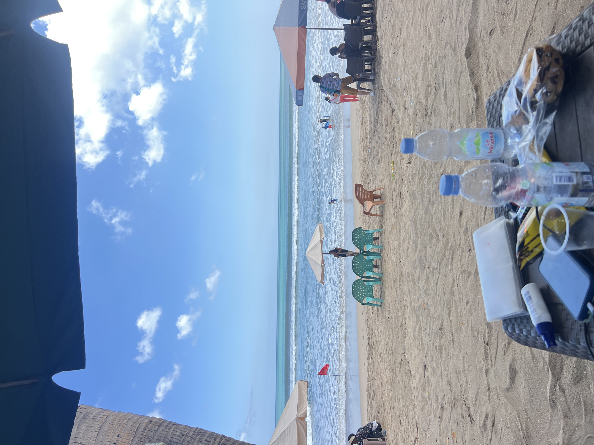 This is a ground-level, eye-level, medium shot of a beach scene in Kuta, Indonesia. The foreground features a wicker table with two plastic water bottles, one of which is labeled "Le Minerale." Also on the table are a white packet, a tube of sunscreen, a smartphone, and a plastic cup with some yellow liquid. The beach stretches out to the ocean, with several beach chairs and umbrellas set up. In the background, several people are visible, some wading in the water and others sitting under umbrellas. The ocean is calm with gentle waves, and the sky is blue with scattered white clouds. A red flag is planted in the sand near the water's edge.