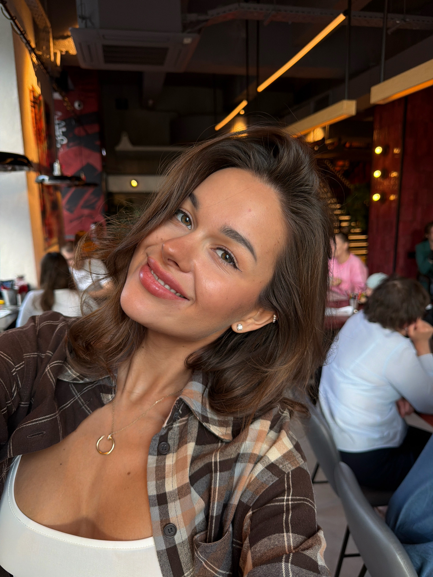 The image is a selfie of a young woman with brown hair, wearing a brown and white plaid shirt and a white tank top. She is wearing a gold necklace with a circular pendant and a diamond stud earring. She is smiling at the camera. In the background, there are other people sitting at tables in what appears to be a restaurant or cafe. The interior features exposed brick walls, industrial-style lighting, and dark red accents. The location is identified as the city of Krasnoyarsk, Russia, based on user context.