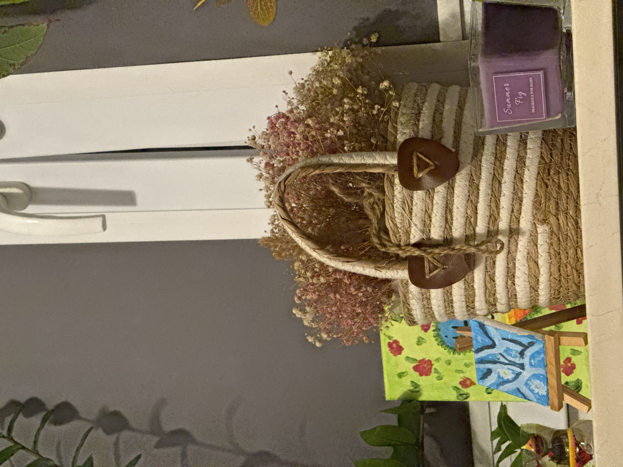 The image depicts a close-up of a windowsill adorned with various decorative items. In the center is a woven straw basket filled with dried flowers, predominantly in shades of pink and beige. The basket has two brown leather handles with a triangular cutout. To the left of the basket, a small canvas painting with a green background and red floral motifs stands upright. In front of this painting, an even smaller canvas is propped on a wooden easel, displaying a blue and white abstract design. To the right of the woven basket, a square glass jar contains a purple candle with a label that reads "Summer Fig FRAGRANCE FOR HOME". Behind these objects, a white window frame is visible against a grey wall. The lighting suggests an indoor setting, possibly in a residential area. The image does not contain any information to suggest a specific location in Łódź, Poland.