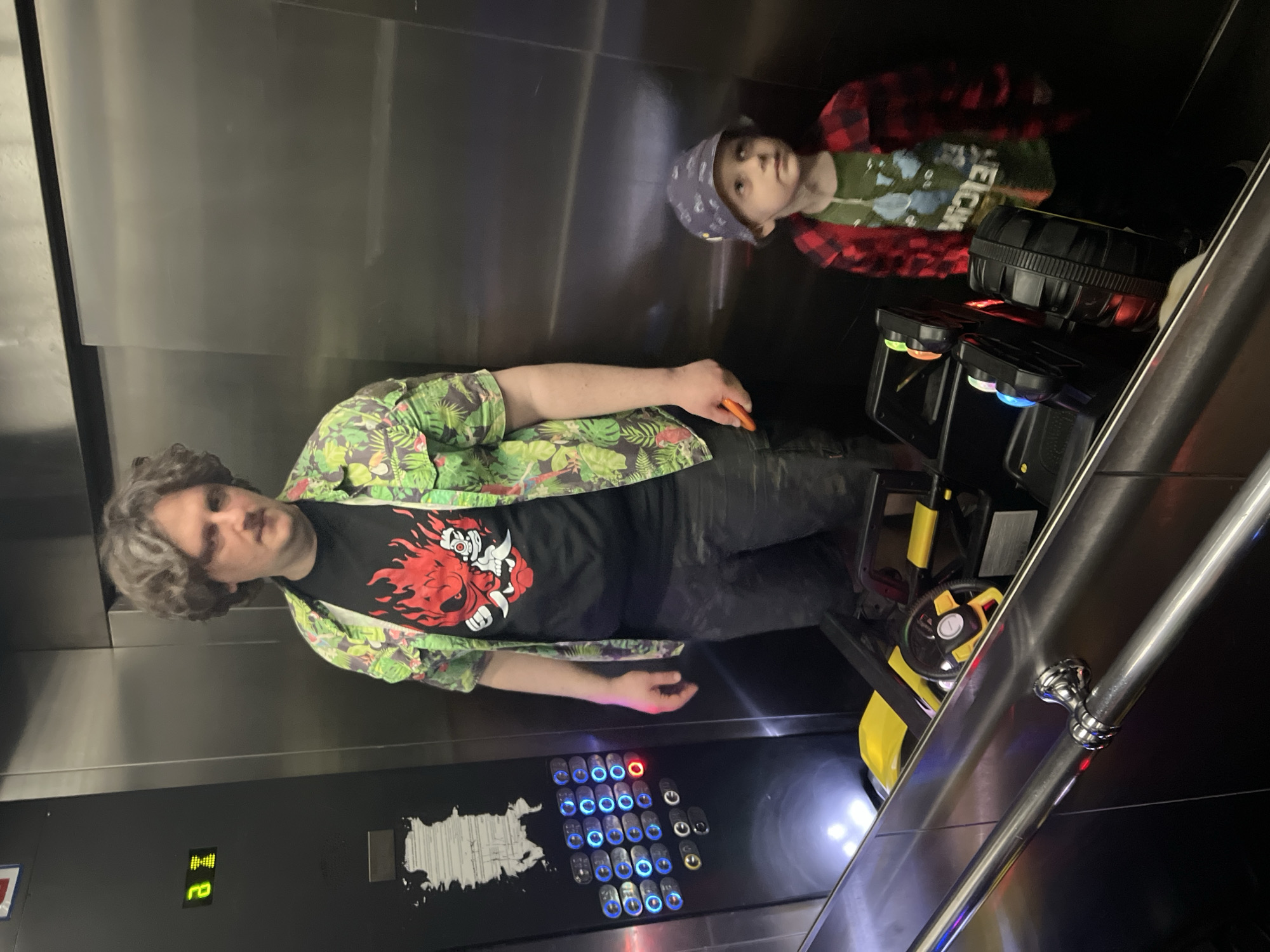 The image shows a man and a child inside an elevator. The man is wearing a green tropical-print shirt over a black T-shirt with a red and white graphic, and dark pants. He is standing near the elevator's control panel, which is to his left and has multiple illuminated blue buttons, one red button, and other gray buttons. The elevator indicator shows the floor number '2' in green. A child wearing a bandana and a red and black plaid shirt, is reflected in a side wall. There's also a toy car-like object with yellow and black components in the elevator. The interior of the elevator appears to be made of dark, reflective materials. There is no information to conclude this is Mytishchi, Russia.