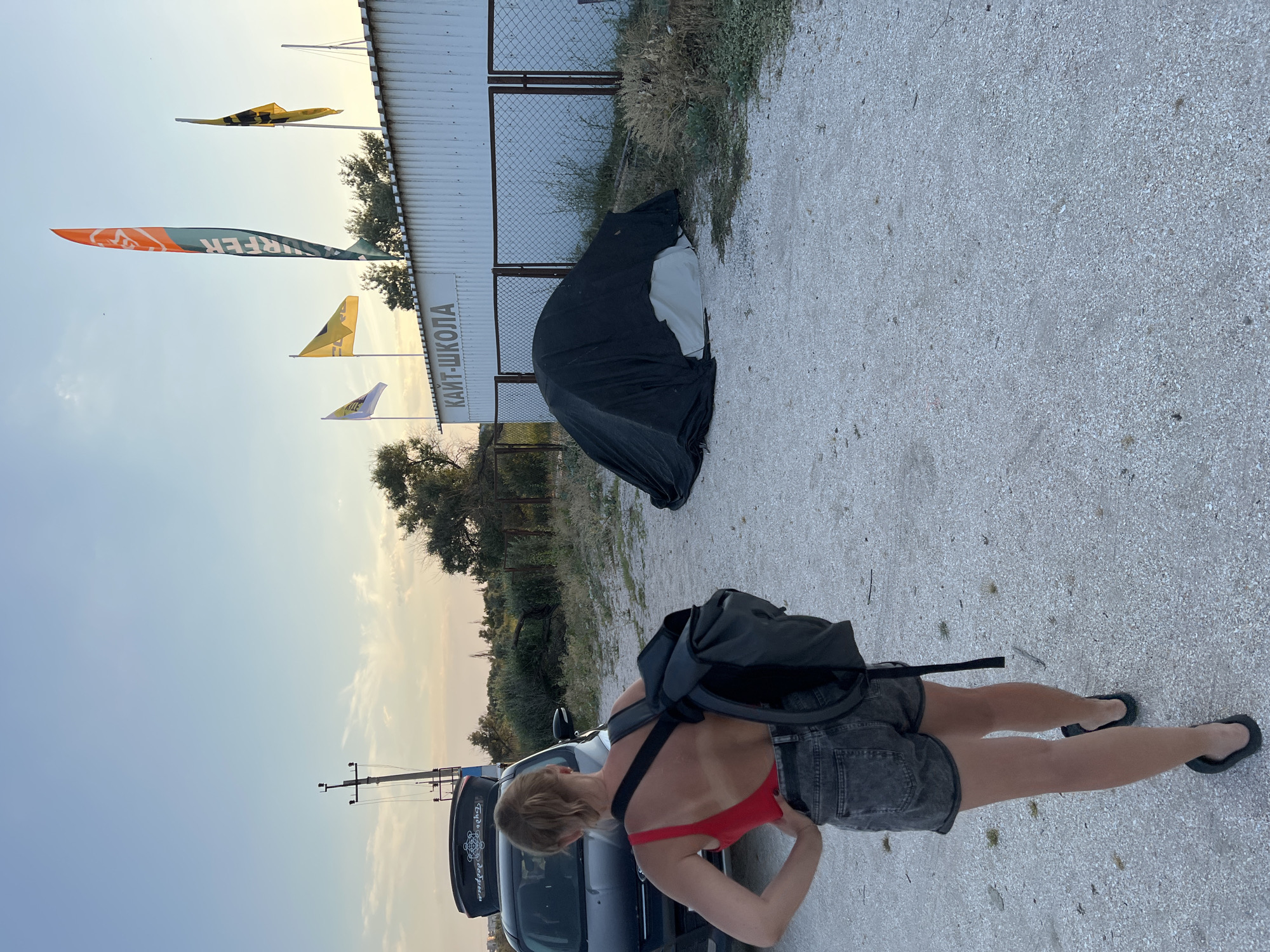 A young woman with blonde hair tied back, wearing a red swimsuit and denim shorts, is standing with her back to the camera. She has a black backpack on. To her left is a dark-colored car with its trunk open, containing a black cargo box. In the background, a white building with "КАЙТ-ШКОЛА" (Kite School) written on it is visible. Several flags are flying in front of the building. A tent covered with a dark tarp is on the ground to the right of the building. The ground is covered with gravel and some vegetation. The scene appears to be outdoors, possibly at dusk, with a clear sky overhead.