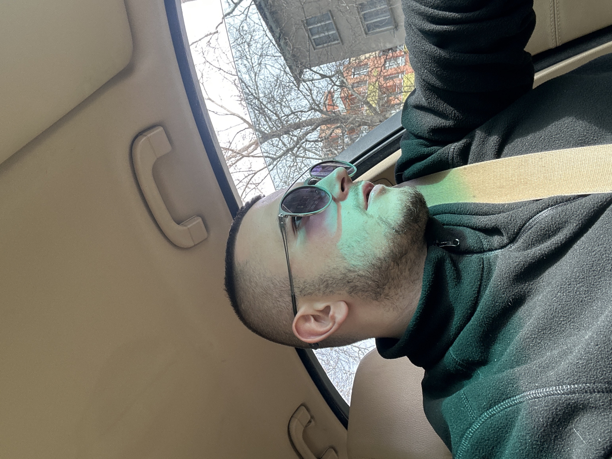A man is seated in a car, wearing a black fleece jacket and sunglasses. A seatbelt is fastened across his chest. He has a bruise around his left eye. Outside the car window, bare tree branches and buildings are visible. The car's interior is tan. The location is Plovdiv, Bulgaria based on the style of the buildings, trees and general ambience.