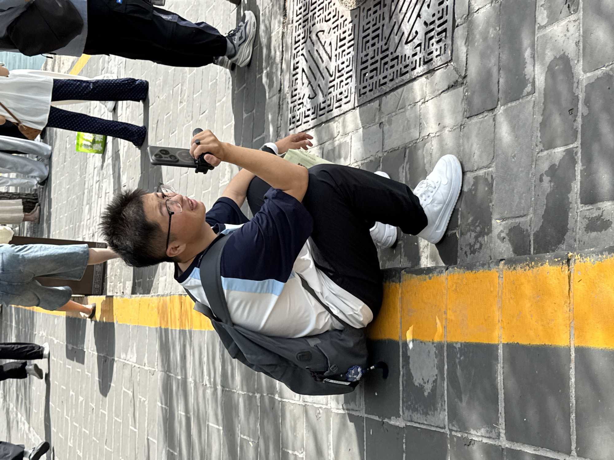 This outdoor image, captured in a pedestrian area in Haidian District, China, features a young East Asian male in the foreground, crouched low next to a prominent yellow line painted on dark gray paving stones. The male, appearing to be in his late teens or early twenties, wears dark-rimmed glasses, a short-sleeved polo shirt with a white torso, dark blue sleeves, and light blue and white horizontal stripes across the chest. He is dressed in black trousers and white low-top sneakers. A dark gray backpack is situated on his back, from which a blue and white ornamental tassel hangs from a side pocket. He holds a dark-colored smartphone vertically, mounted on a black handheld grip, directing it towards his right. His mouth is slightly ajar, and he appears to be actively engaged with what he is recording or observing.
The ground consists of regularly laid dark gray square paving stones, marked by a bright, weathered yellow line running vertically through the lower left and center of the frame. To the right of the main subject, a dark metal grate with a geometric pattern is embedded in the pavement. In the midground and background, several other individuals are partially visible, suggesting a public space with pedestrian traffic. These include a person in dark denim shorts or capris, another wearing dark blue trousers with a repetitive pattern, and others in light-colored attire. A light green bag is visible on the pavement. The scene is illuminated by strong sunlight, casting distinct shadows from the people and structures, indicating a clear day.