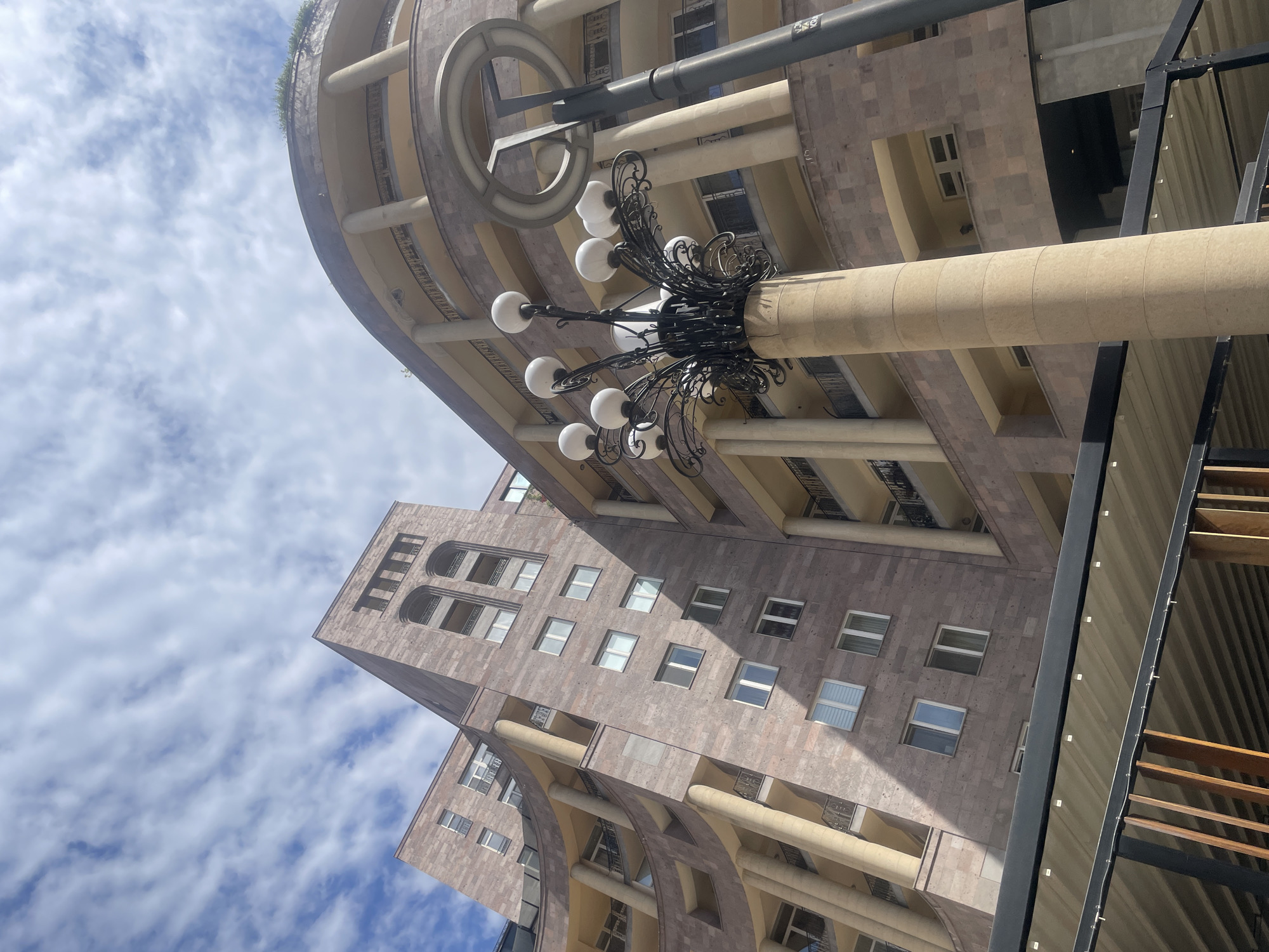 The image shows architecture in Yerevan, Armenia. The buildings are constructed of light brown stone. The building on the right has curving floors with columns. There are rectangular windows. There is an ornate lamp in the foreground with round white bulbs. There is a blue sky with puffy white clouds.