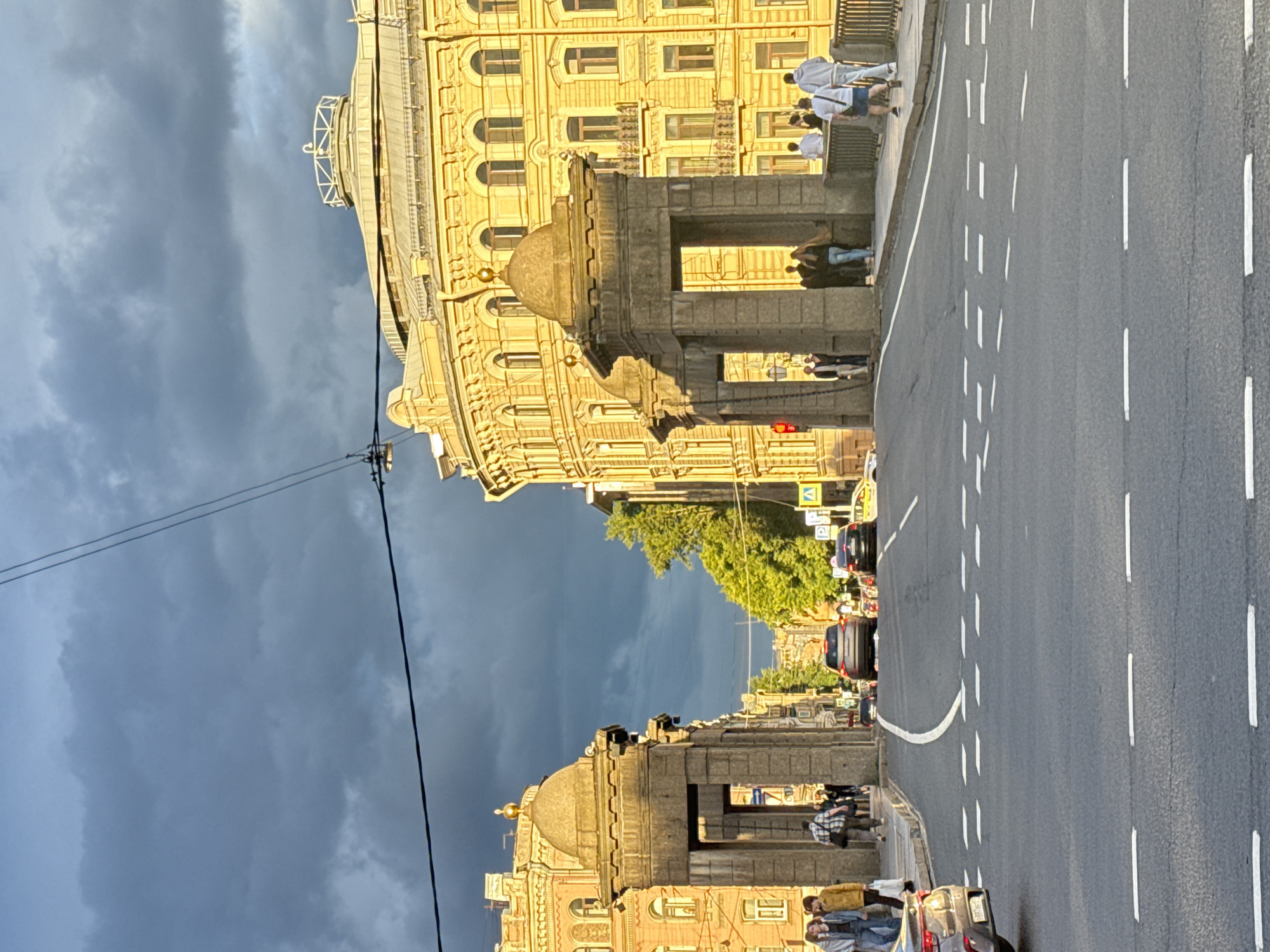 The image depicts a street scene in Saint Petersburg, Russia, likely during the late afternoon or early evening, given the warm, golden light illuminating the ornate yellow building on the left. Dark, dramatic clouds fill the sky, suggesting an approaching storm or the remnants of one. The street is paved with asphalt and marked with white lane dividers. Several cars are visible, suggesting moderate traffic.
On the sidewalk to the left of the road, architectural elements of an old building are prominent, featuring arched windows and decorative stonework. A grand archway, adorned with a dome-like structure, stands out. People are walking along the sidewalk, some in groups, interacting with each other or simply passing by. Their clothing suggests casual attire, and their postures do not convey any strong emotions, appearing to be engaged in everyday activities.
Further down the street, more buildings and trees are visible, hinting at a well-established urban environment. A traffic light is visible on the right side of the road, indicating an intersection or crossing. The presence of power lines crossing the frame also adds to the urban context. Overall, the scene captures a moment of urban life under a dynamic sky, with the historical architecture of Saint Petersburg providing a grand backdrop.