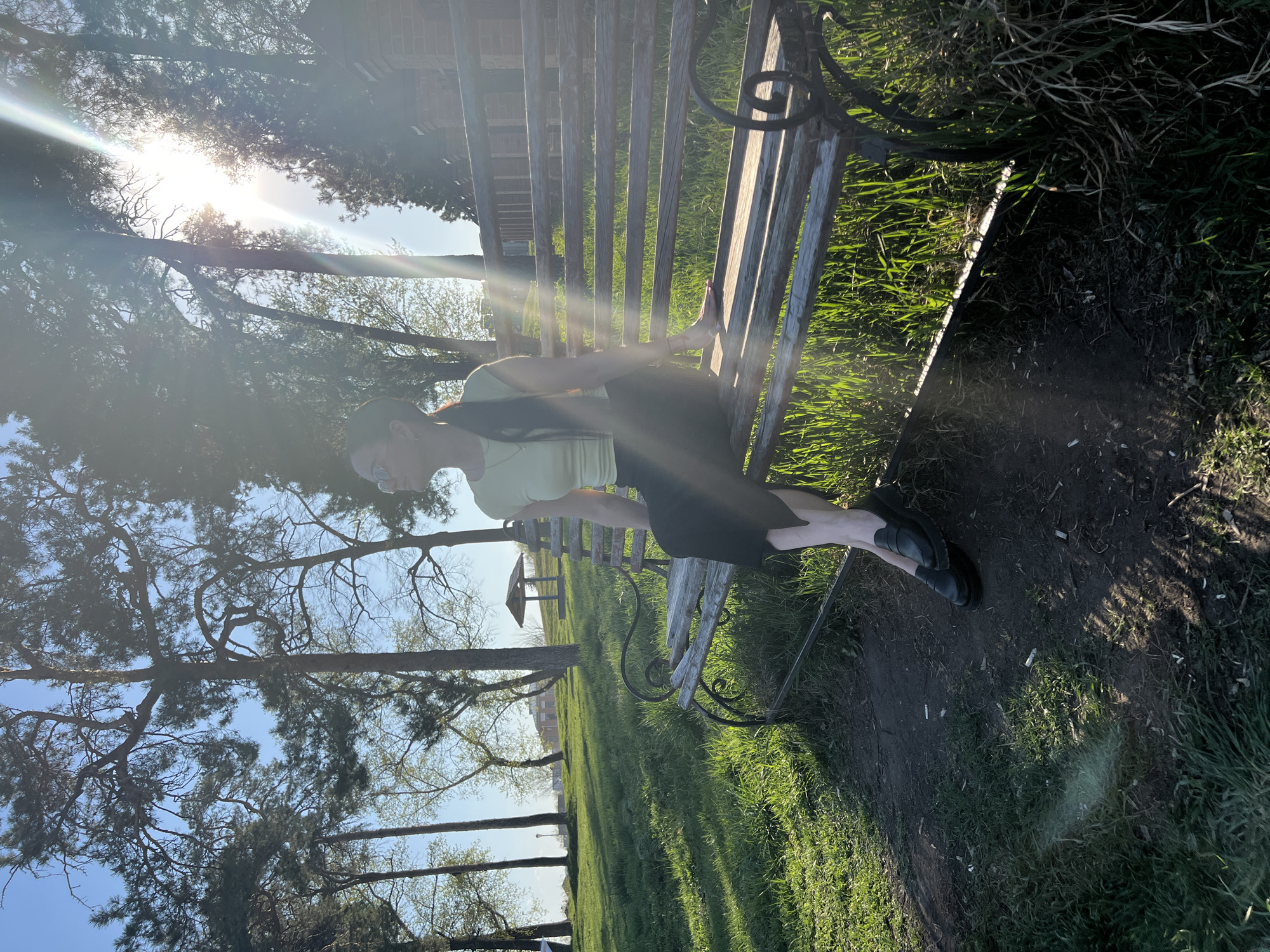 A person is sitting on a weathered wooden and metal bench in a park-like setting. The individual is wearing a light-colored top, a dark skirt, and dark shoes. Light streams through the trees and reflects off the bench. The surrounding environment features grassy areas, trees, and possibly another park structure in the background. The location is in Sviyazhsk, Russia.