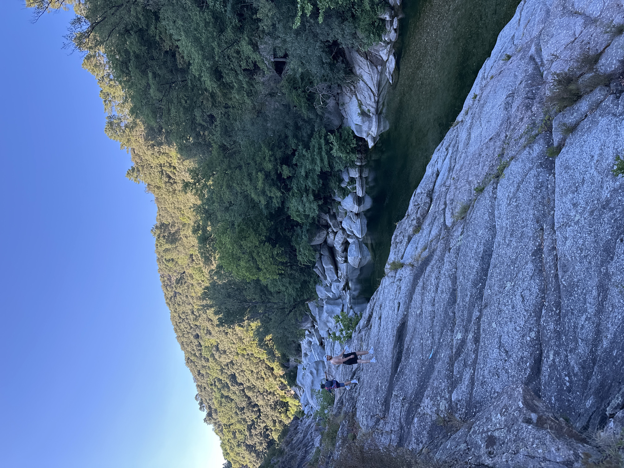 The image depicts a scenic view of the Gardon River in Anduze, France. The river flows through a rocky gorge lined with lush green trees and vegetation on the hillside. The water is clear and calm, reflecting the surrounding landscape. In the foreground, two individuals are standing on the rocky terrain. One person is wearing a dark backpack and shorts, while the other is wearing black shorts and a white cap. They appear to be casually observing the river or preparing to engage in an activity. The overall scene is serene and natural, highlighting the beauty of the French countryside.