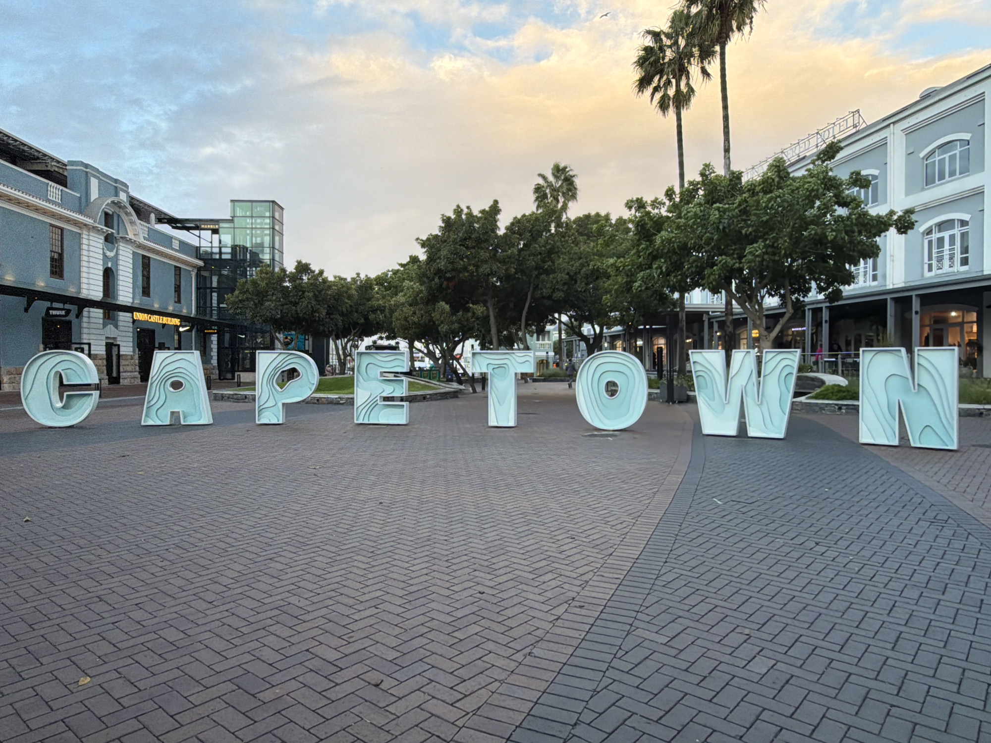 This image captures a wide shot of the iconic "CAPETOWN" sign in an urban outdoor setting. The sign itself consists of large, individual block letters, light teal or mint green in color, featuring a layered, topographic map-like pattern on their surfaces. They are arranged in a row across a paved ground, spelling out "CAPETOWN".
The scene appears to be a public square or promenade, paved with dark gray interlocking bricks laid in a herringbone pattern. In the background, there are multi-story buildings, characterized by light blue-grey facades with white trim and arched windows, giving them a classic architectural feel. On the left side, a more modern glass and steel structure is integrated with one of these buildings. Lush green trees are scattered throughout the scene, and several tall palm trees are visible, especially on the right side and in the center-left, reaching towards the sky.
The sky is partly cloudy with soft, warm hues of yellow and orange mixing with cooler blues, suggesting either late afternoon or early morning. The lighting is diffuse and gentle.
Visible text includes the prominent "CAPETOWN" letters. On the buildings in the background, shop signs or building names can be identified: "THULE" is visible on a sign to the left, and "UNION CASTLE BUILDING" is displayed on another sign. A "NIKE" logo is partially visible on a storefront, and the word "MARBLE" can also be seen. No specific human activity is central, but the arrangement of the letters implies a landmark designed for visitors and photography. The overall atmosphere is peaceful and inviting.