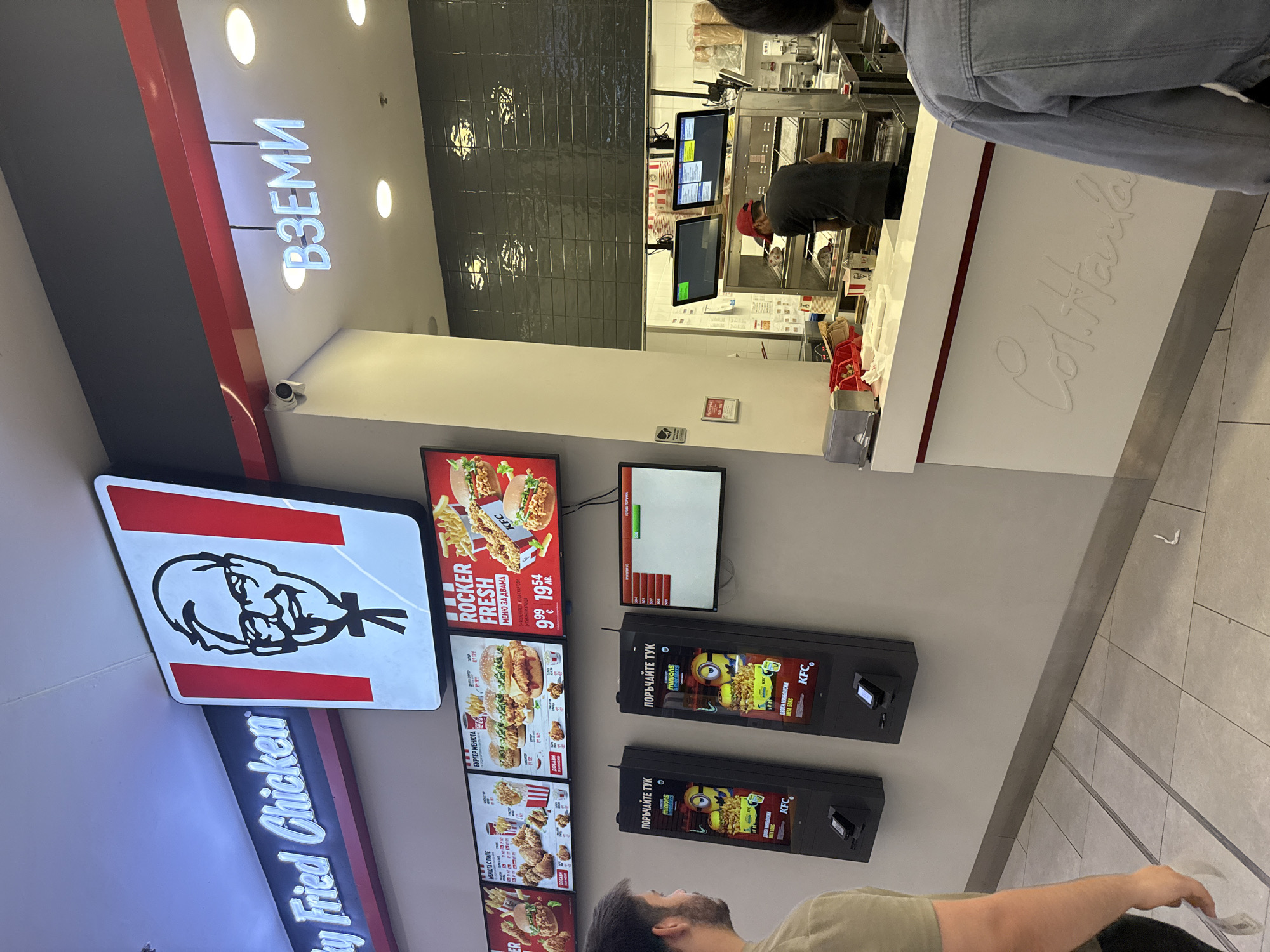 The image shows the interior of a KFC restaurant, likely in Stara Zagora, Bulgaria, given the Cyrillic text on the menu boards and signage. A customer is standing in front of two self-order kiosks. The kiosks display promotions for KFC meals, including "Minyons" themed boxes. Above the kiosks, large digital menu boards display various KFC food items and prices. To the right, a counter area is visible with employees preparing food behind it. A small screen on the wall displays order numbers. The overall environment appears to be a modern fast-food establishment with typical interior decor and branding. The sign "ВЗЕМИ" (Take) is visible above the counter. The floor is tiled and the walls are a neutral color.