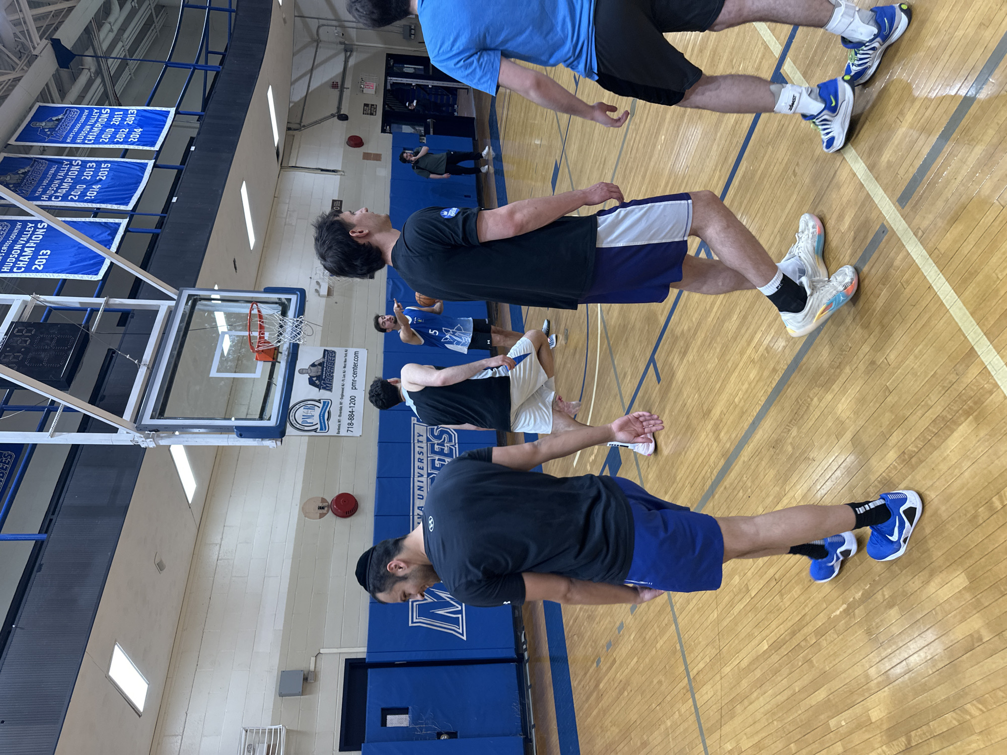 This image captures an indoor scene within a gymnasium, likely in New York, USA, depicting several young men on a basketball court. The setting features a polished wooden floor with court markings, basketball hoops with backboards and nets, and blue padded walls. Fluorescent lighting illuminates the space.
The subjects are approximately seven men, appearing to be athletes, dressed in various athletic attire including t-shirts, shorts, and basketball shoes. They are mostly standing, some with focused or neutral expressions, suggesting they are engaged in a warm-up, practice, or listening to instructions. One individual in the background wears a blue jersey with the number "5", and another man near the center holds a basketball. Their postures indicate a state of readiness or light activity.
Notable details include several blue and white championship banners hanging above the court. These banners display text such as "MACCABEES," "MEN'S CROSS COUNTRY HUDSON VALLEY CHAMPIONS 2013," "MEN'S VOLLEYBALL HUDSON VALLEY CHAMPIONS 2010 2013 2014 2015," and another "MEN'S CROSS COUNTRY HUDSON VALLEY CHAMPIONS 2010 2011 2012 2013 2014." A sign affixed to the basketball hoop support reads "PMR MACCABEES," including a phone number "718-884-1200" and "pmr-center.com." Partially visible text also shows "VA UNIVERSITY," likely referring to Yeshiva University. A scoreboard is partially visible on the left, displaying "18:88." The overall scene suggests an athletic team engaging in practice or a pre-game routine.